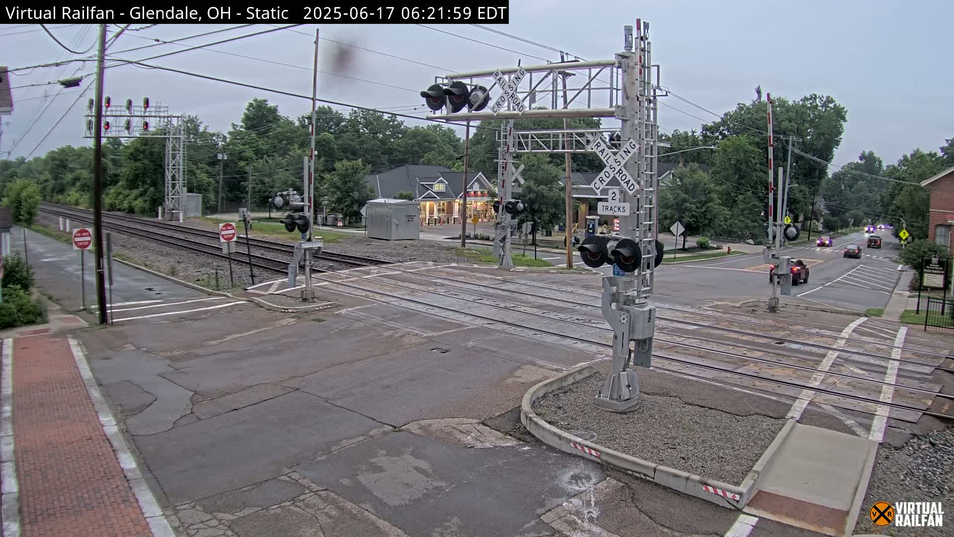 A railroad crossing with lowered gates shows a street intersection under an overcast sky, with several cars stopped and a few others driving in the distance.