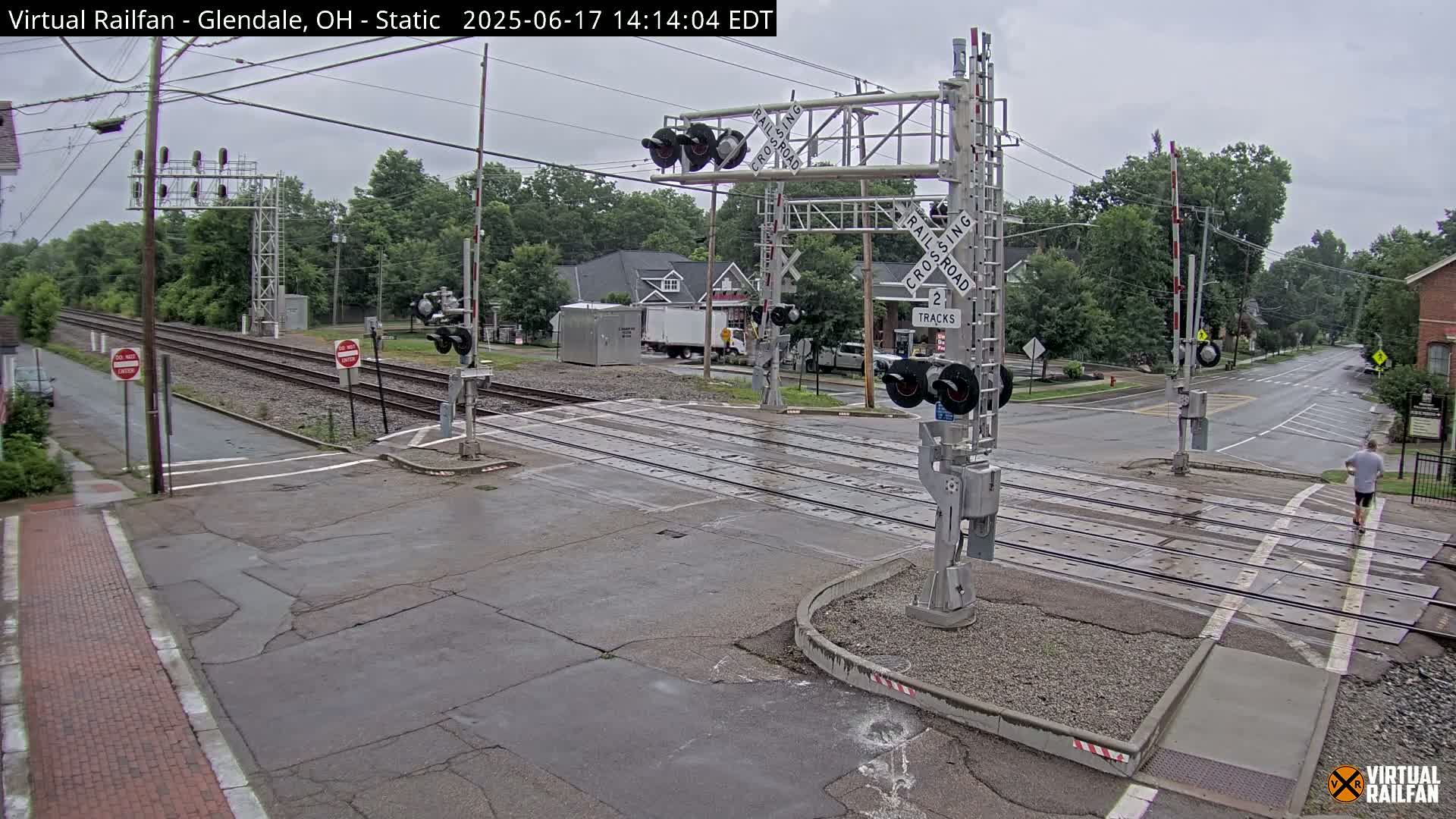 A railroad crossing with lowered gates is shown on an overcast day, with a pedestrian crossing the street in the background.