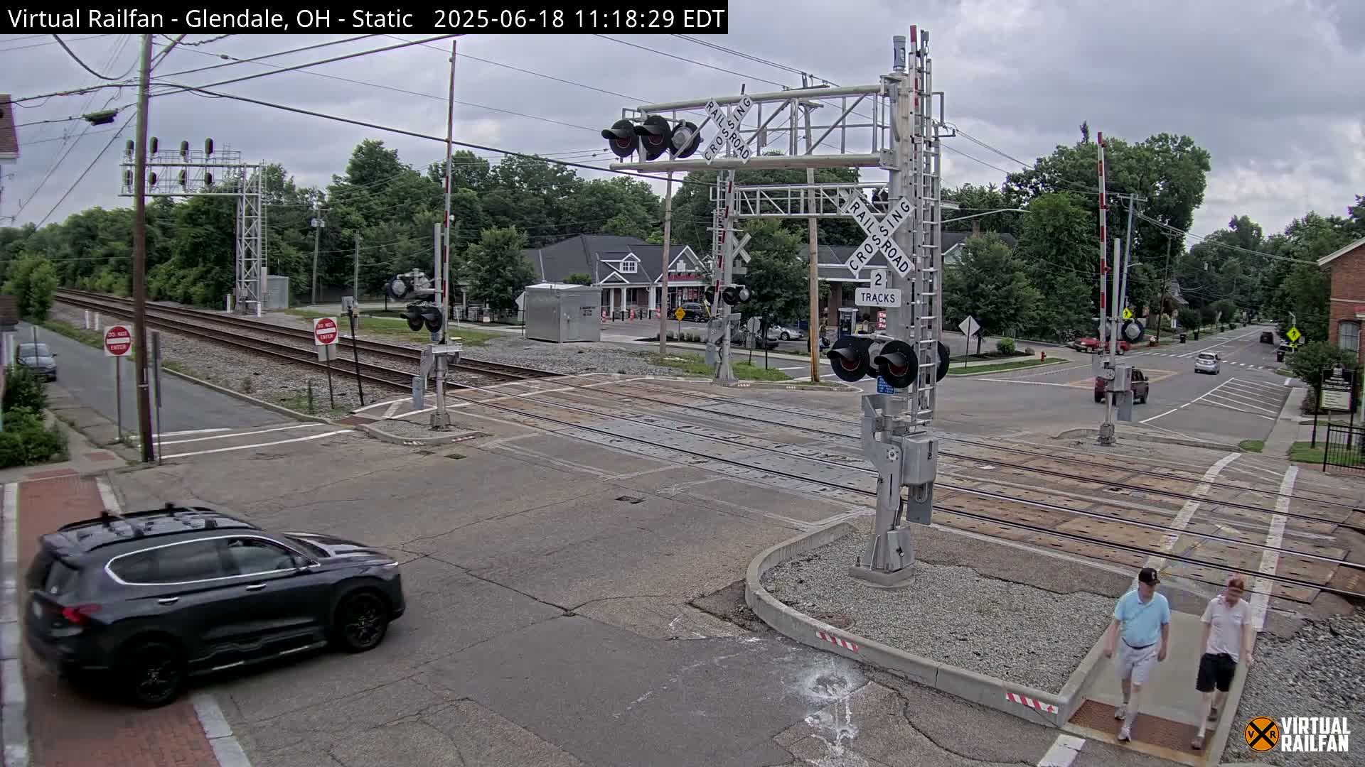 A dark-grey SUV drives past a railroad crossing with two tracks under an overcast sky.