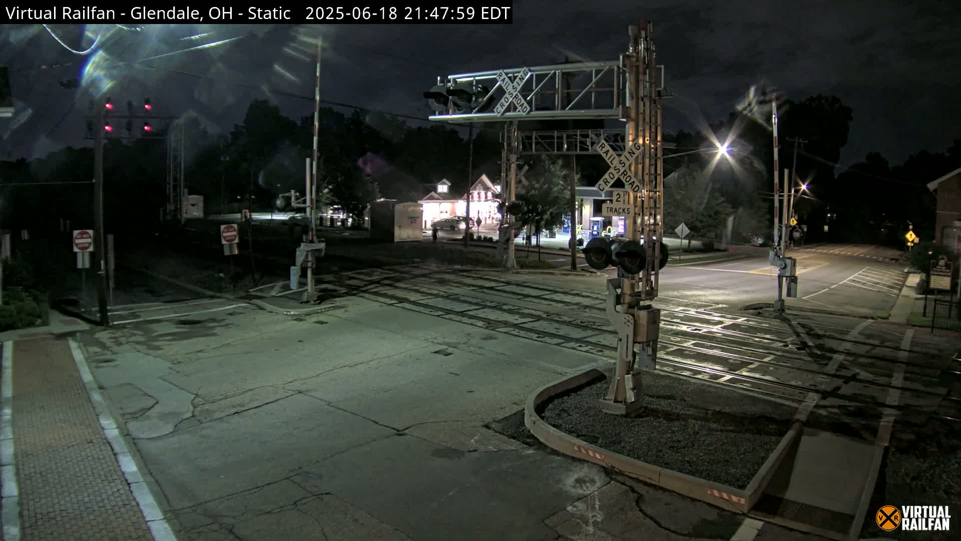 At night, a railroad crossing with lowered gates is illuminated by streetlights and shows two sets of tracks.