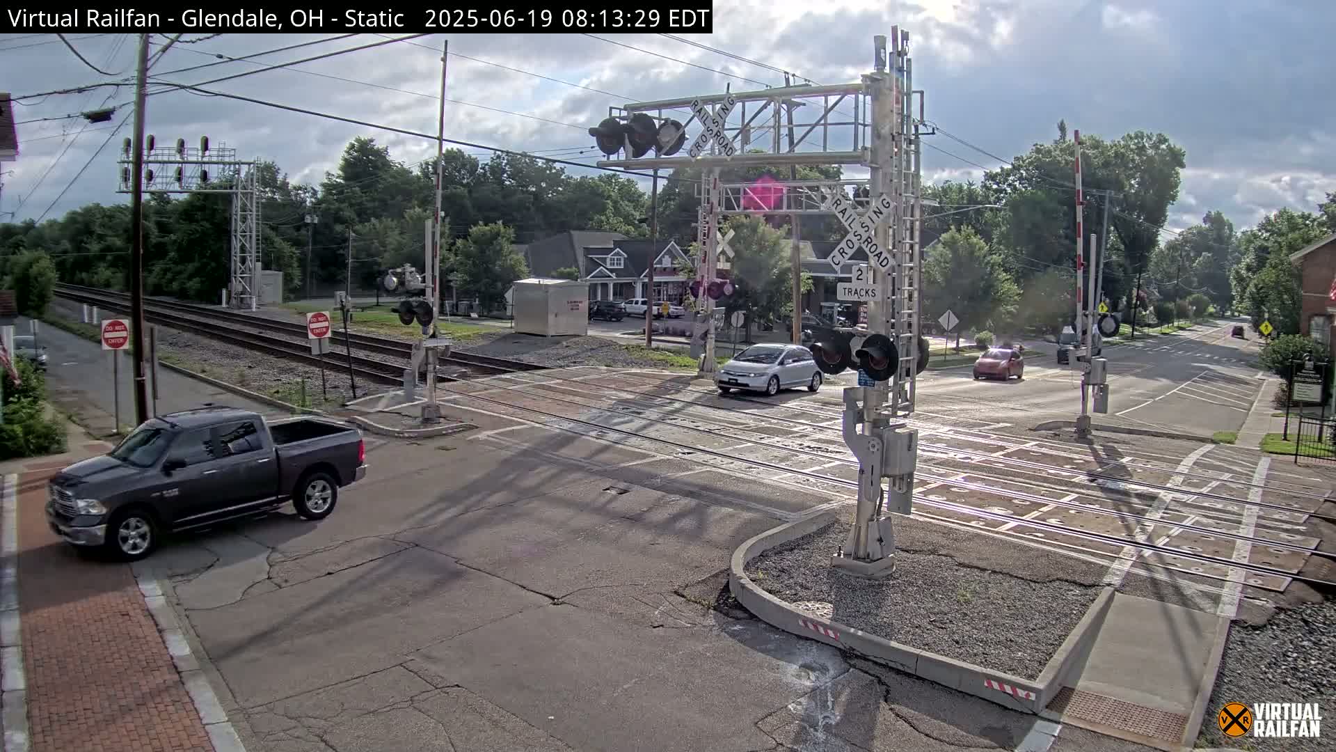 A mostly sunny day shows a railroad crossing with a gray pickup truck and several cars at the intersection, and trees lining the road.