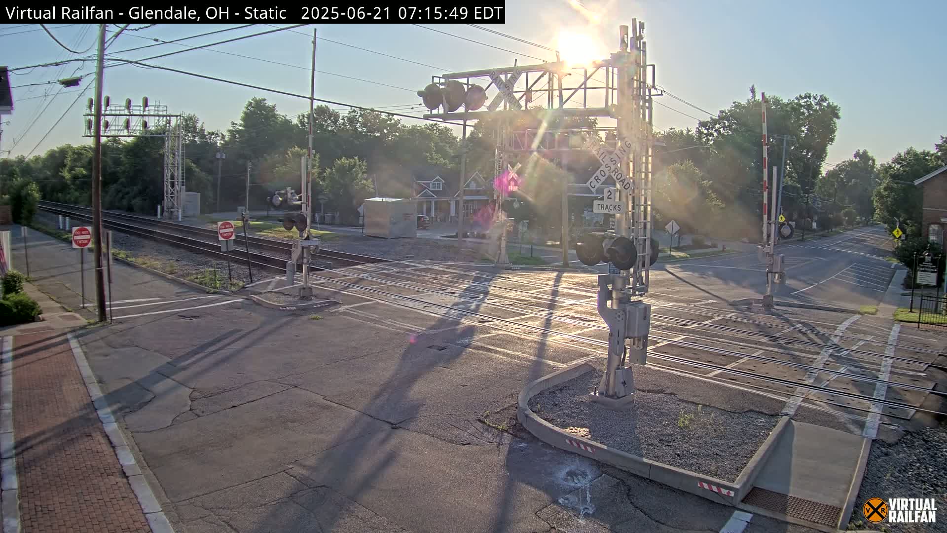 A sunny morning view of a railroad crossing with lowered gates, showing two tracks intersecting a road in a residential area.