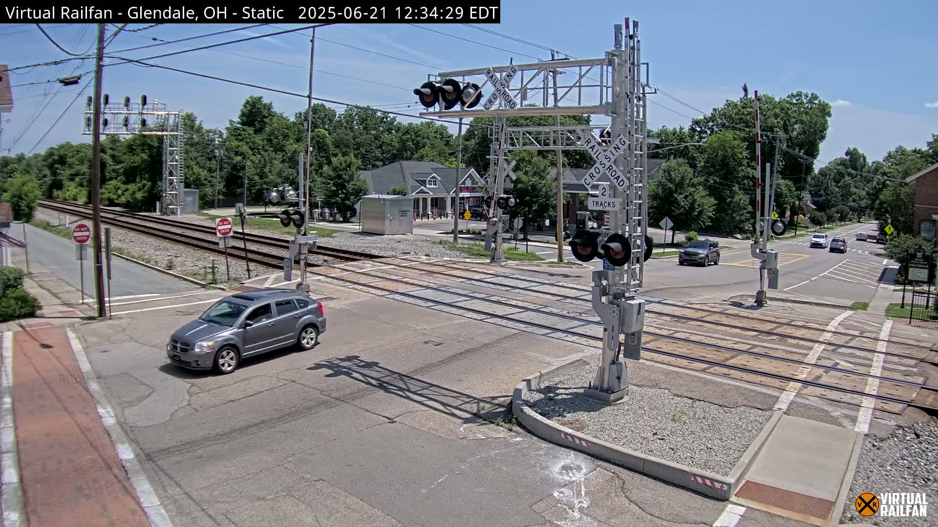 A gray SUV drives across a railroad crossing under a sunny sky, passing through a street intersection near buildings and trees.