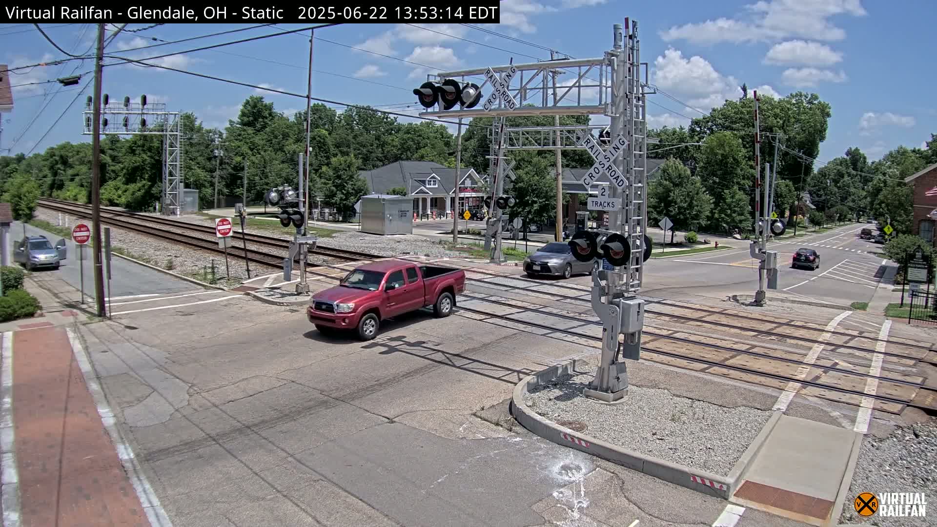 A red pickup truck and several cars are at a railroad crossing under a mostly sunny sky.