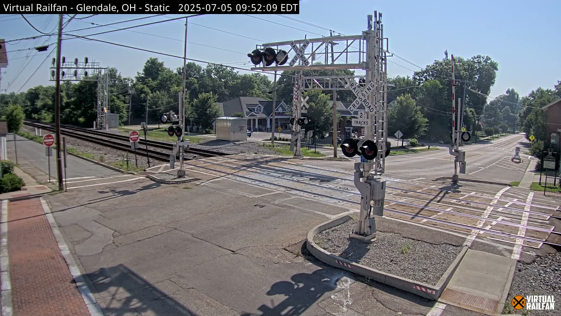 A railroad crossing with two tracks is shown under a clear, sunny sky; the crossing is at the intersection of two roads, with traffic signals and gates in place.