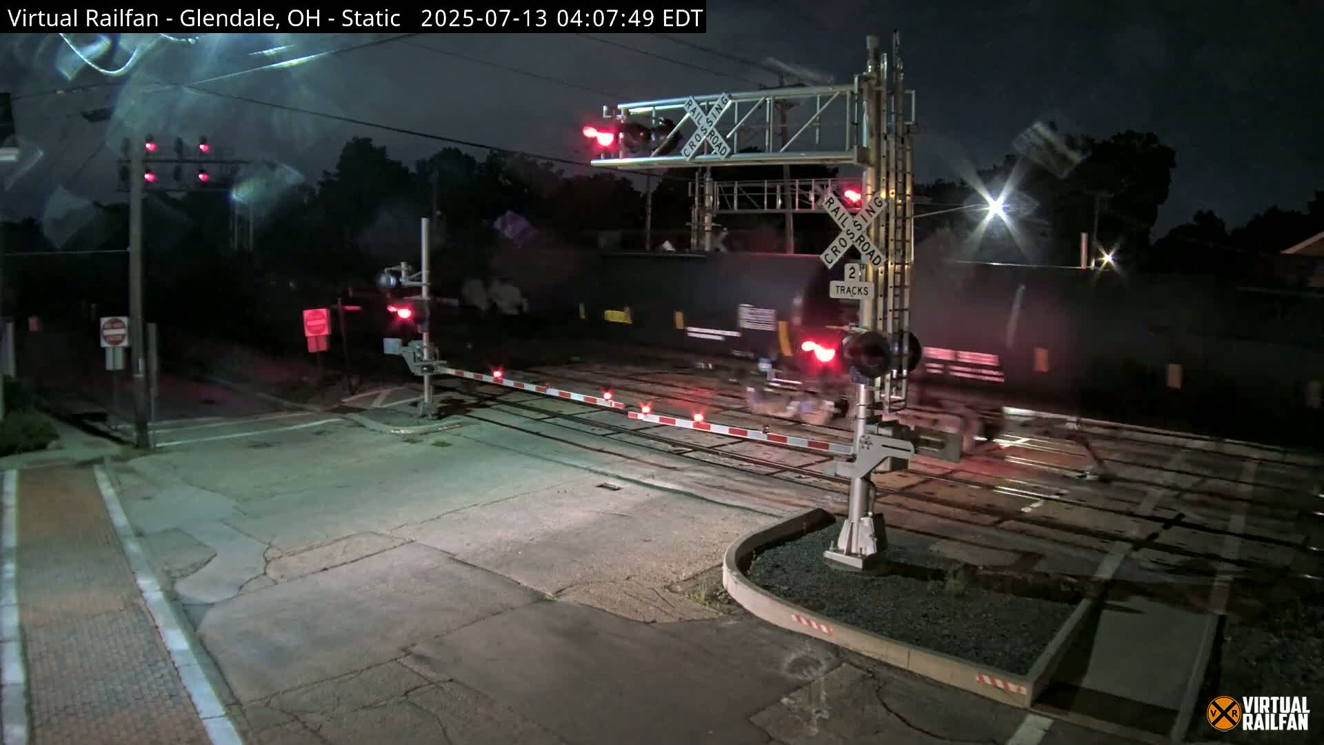 A long freight train passes through a nighttime railroad crossing with its crossing gates down and red lights flashing.