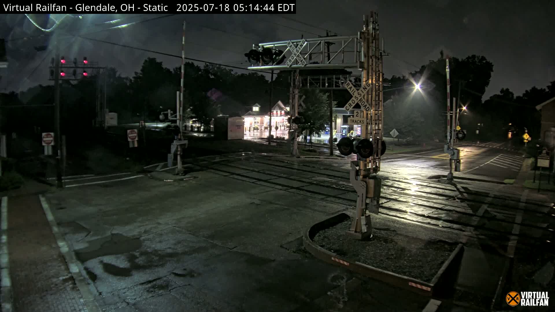 A wet, nighttime view of a railroad crossing with its gates down and red signal lights illuminated.