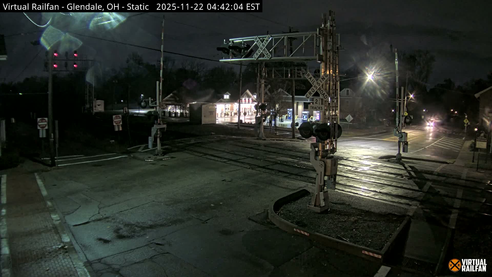 A night scene at a railroad crossing shows multiple tracks, active red signal lights, and raised barriers on wet roads reflecting streetlights and distant vehicle headlights, under misty or light rain conditions.