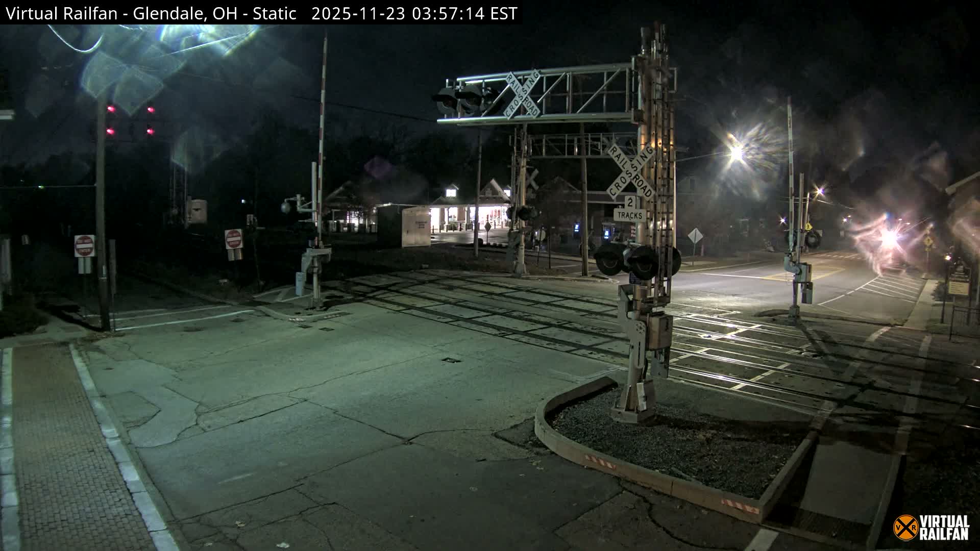 A misty night view captures a street-level railroad crossing with multiple tracks, red warning lights, and illuminated buildings and streetlights in the background.