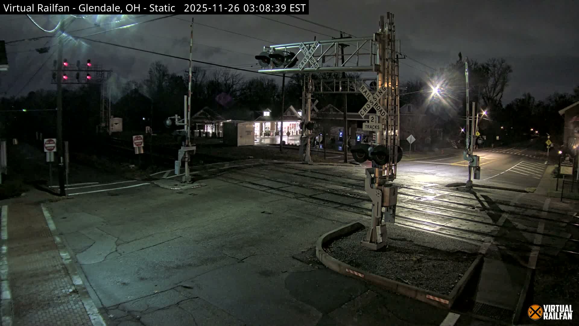 A dark, cloudy night view captures an illuminated railroad crossing featuring multiple tracks, prominent crossing signals with their arms raised, intersecting roads, and softly lit buildings in the background.