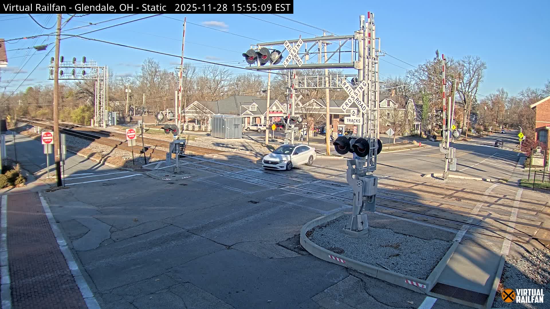 A white car is crossing a railroad intersection with active crossing gates and signals under a clear, sunny sky in a small town setting.