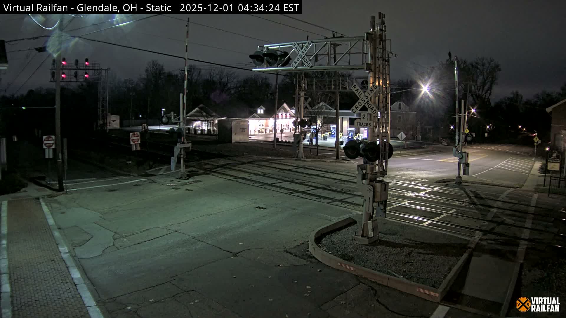 This nighttime image captures an empty railroad crossing with two tracks traversing a paved road, flanked by railway signals with flashing red lights and illuminated by streetlights and distant building lights under a clear sky.