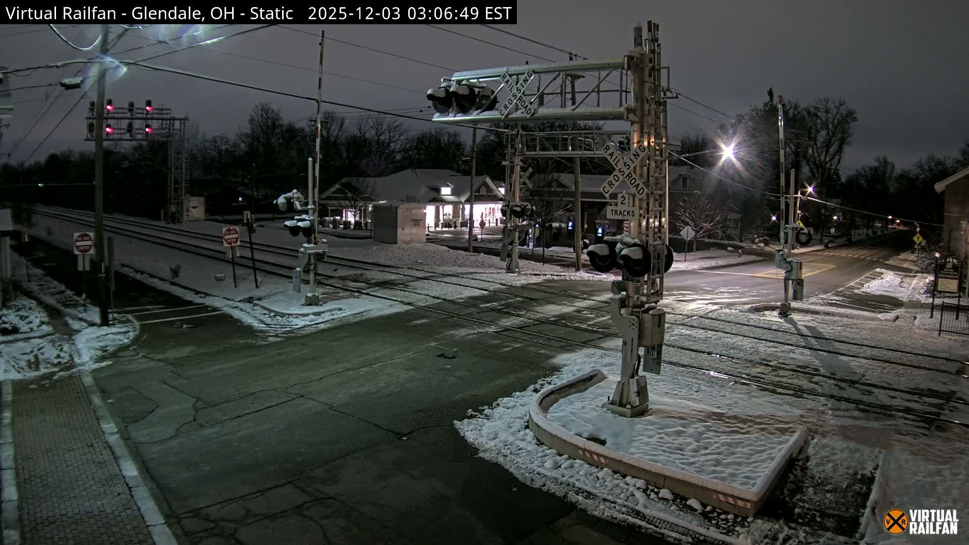 An outdoor scene at night depicts a snowy railroad crossing with tracks and signals covered in fresh snow, an empty intersection, and softly lit buildings in the background under a dark sky.