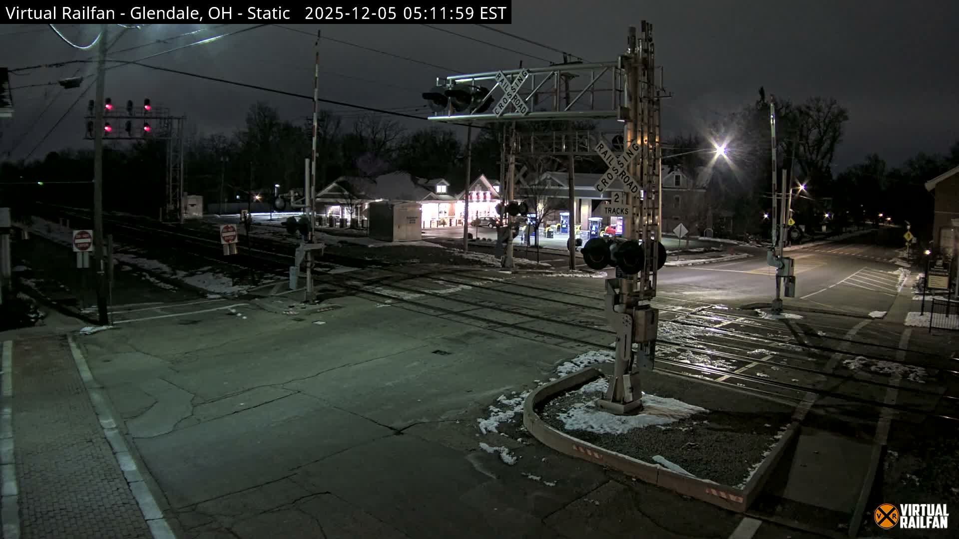 A quiet, wintry night scene at a railroad crossing shows patches of snow on the ground around multiple tracks, illuminated by streetlights and nearby commercial buildings, under a dark, overcast sky.