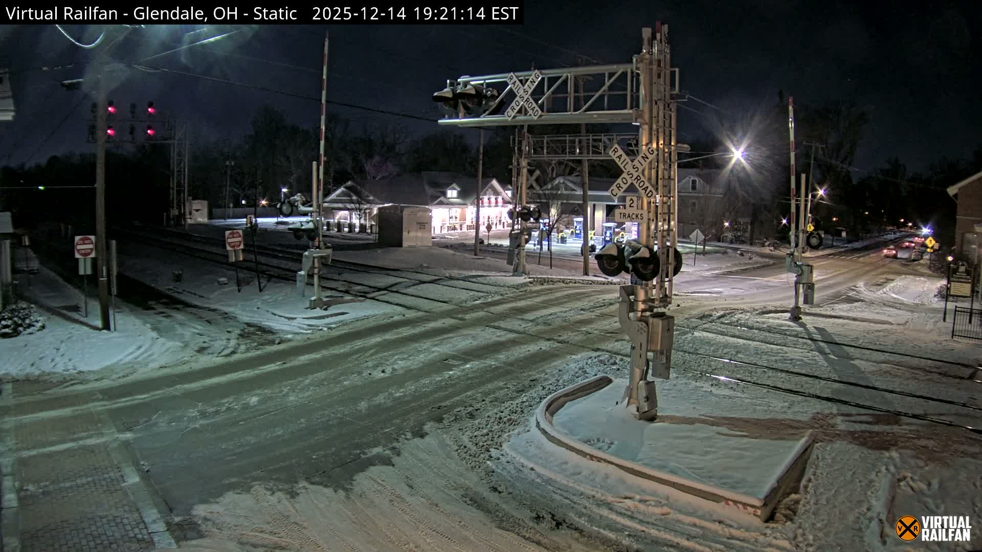 A quiet, wintry night scene at a railroad crossing shows patches of snow on the ground around multiple tracks, illuminated by streetlights and nearby commercial buildings, under a dark, overcast sky.