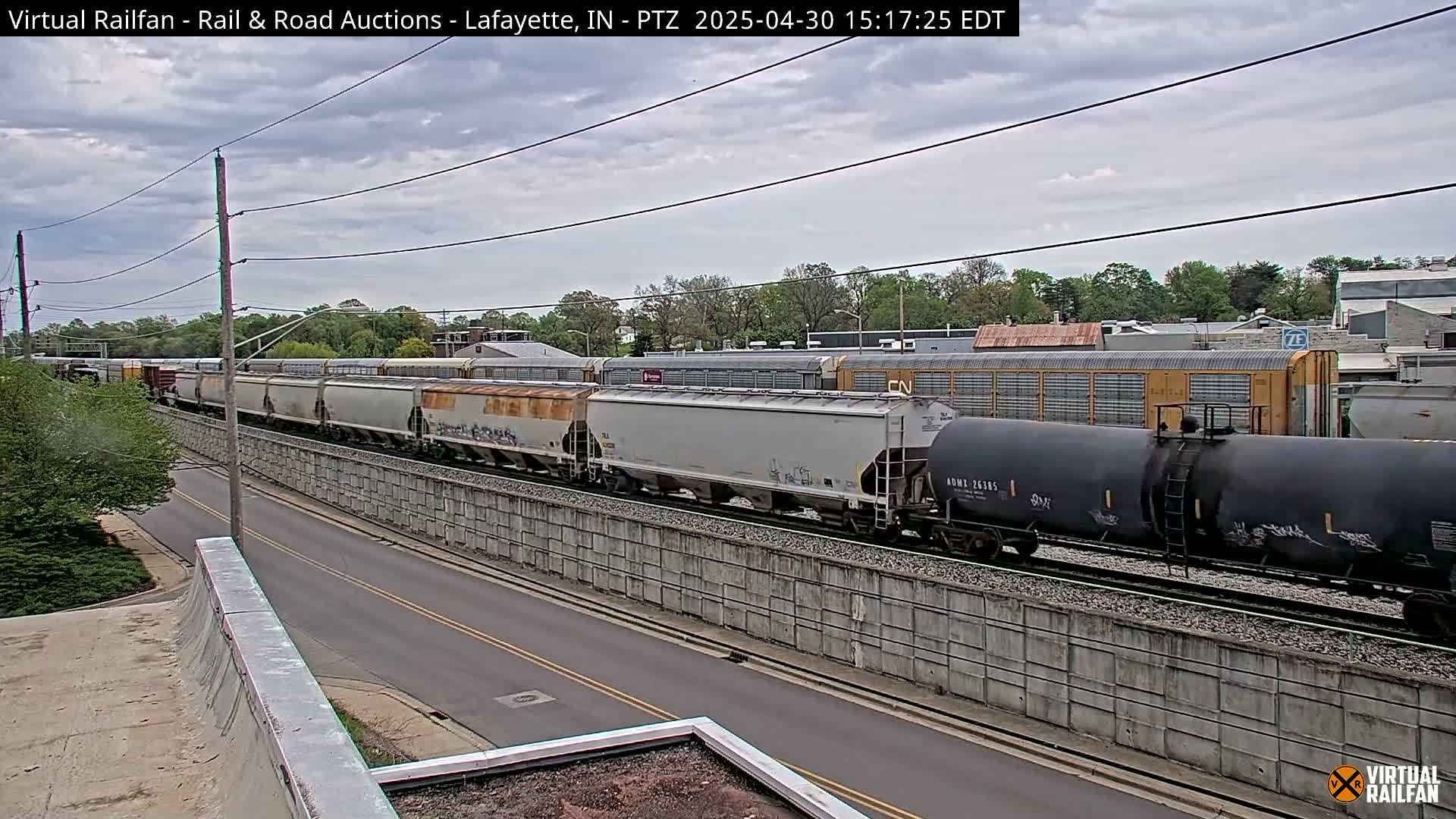 A long freight train passes under an overcast sky alongside a road and a concrete wall.