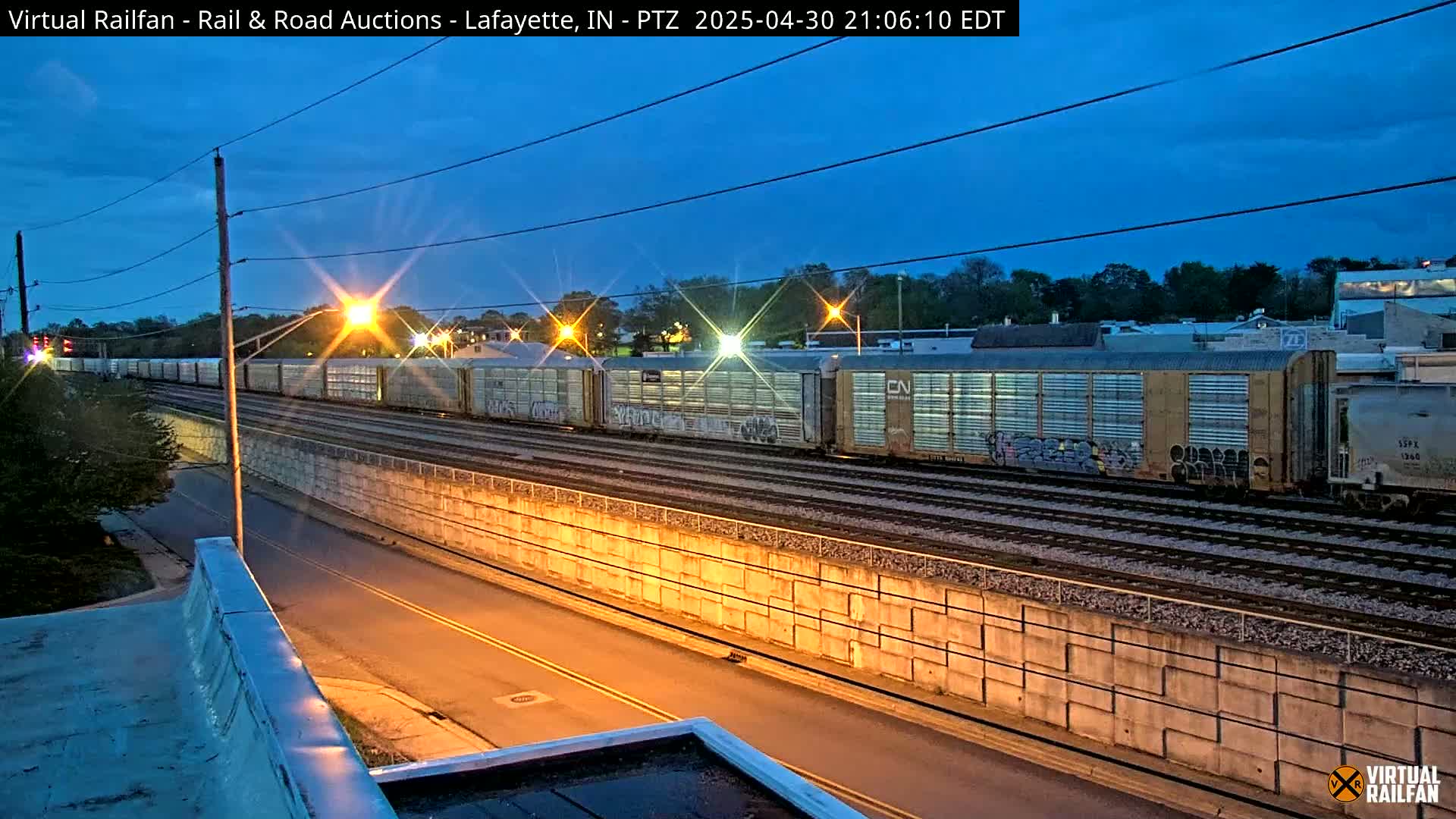 A long freight train passes under a street at twilight, beside a concrete retaining wall.