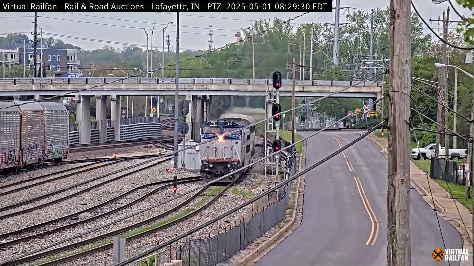 A train locomotive with sparks emitting from its front passes under an overpass on an overcast day.
