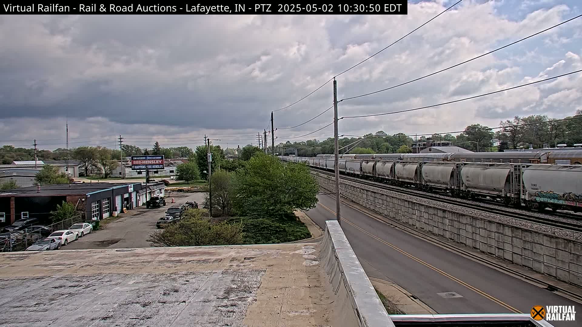 A long freight train passes by under an overcast sky, next to a road and various buildings.