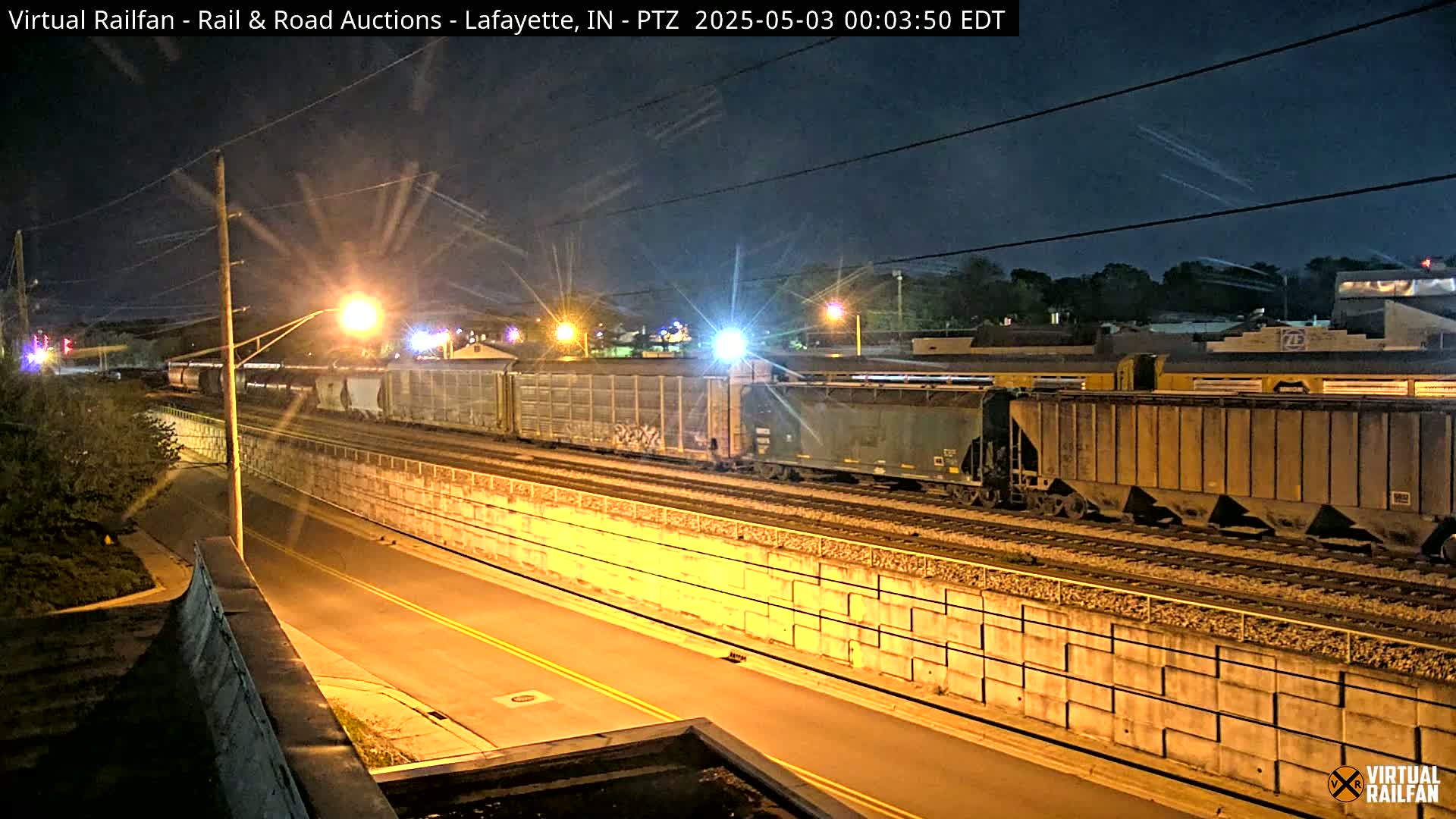 At night, a long freight train passes by a roadway beside a high retaining wall under a dark, clear sky.