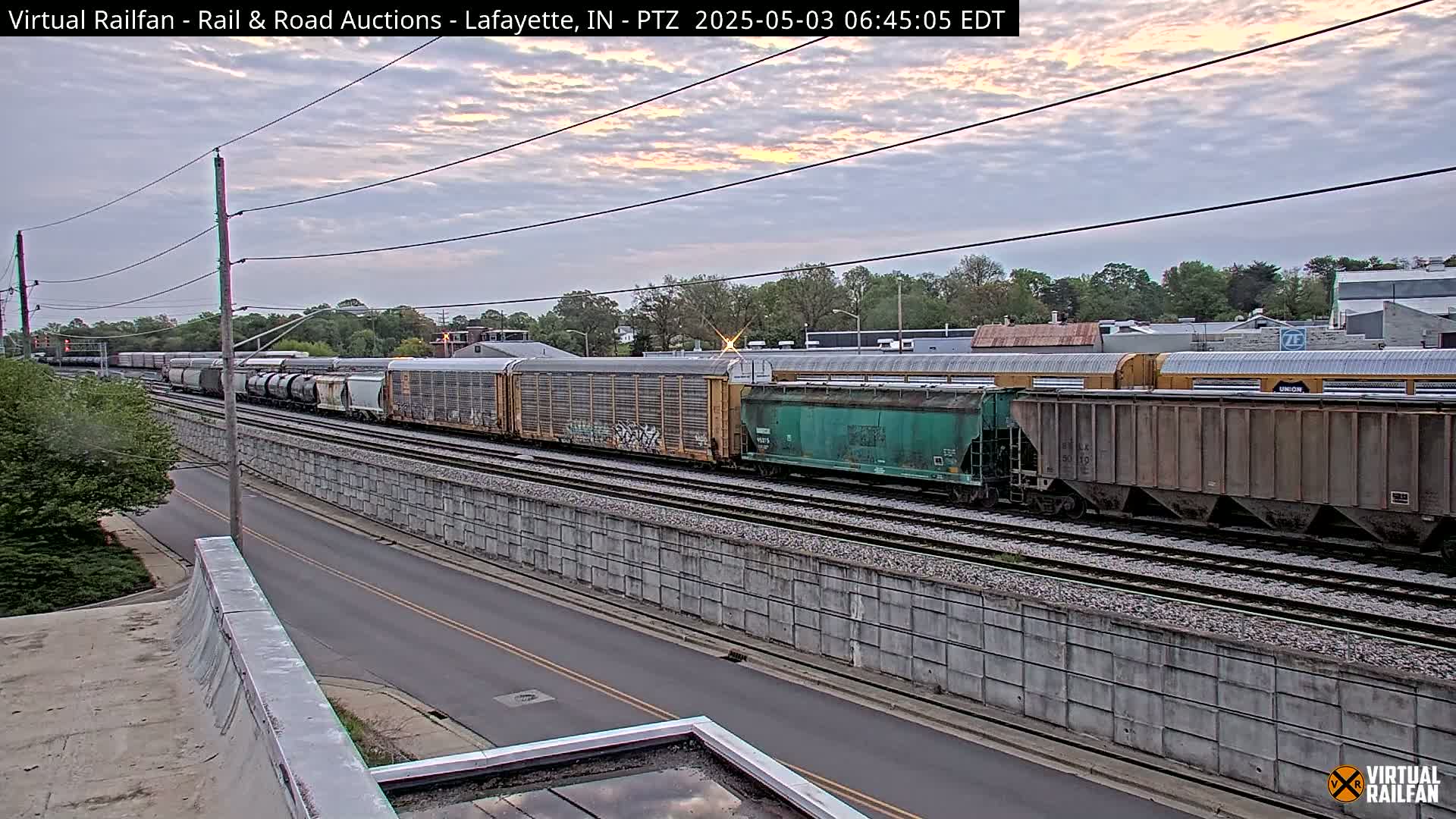 A long freight train passes under an overcast sky, next to a road separated by a concrete barrier.