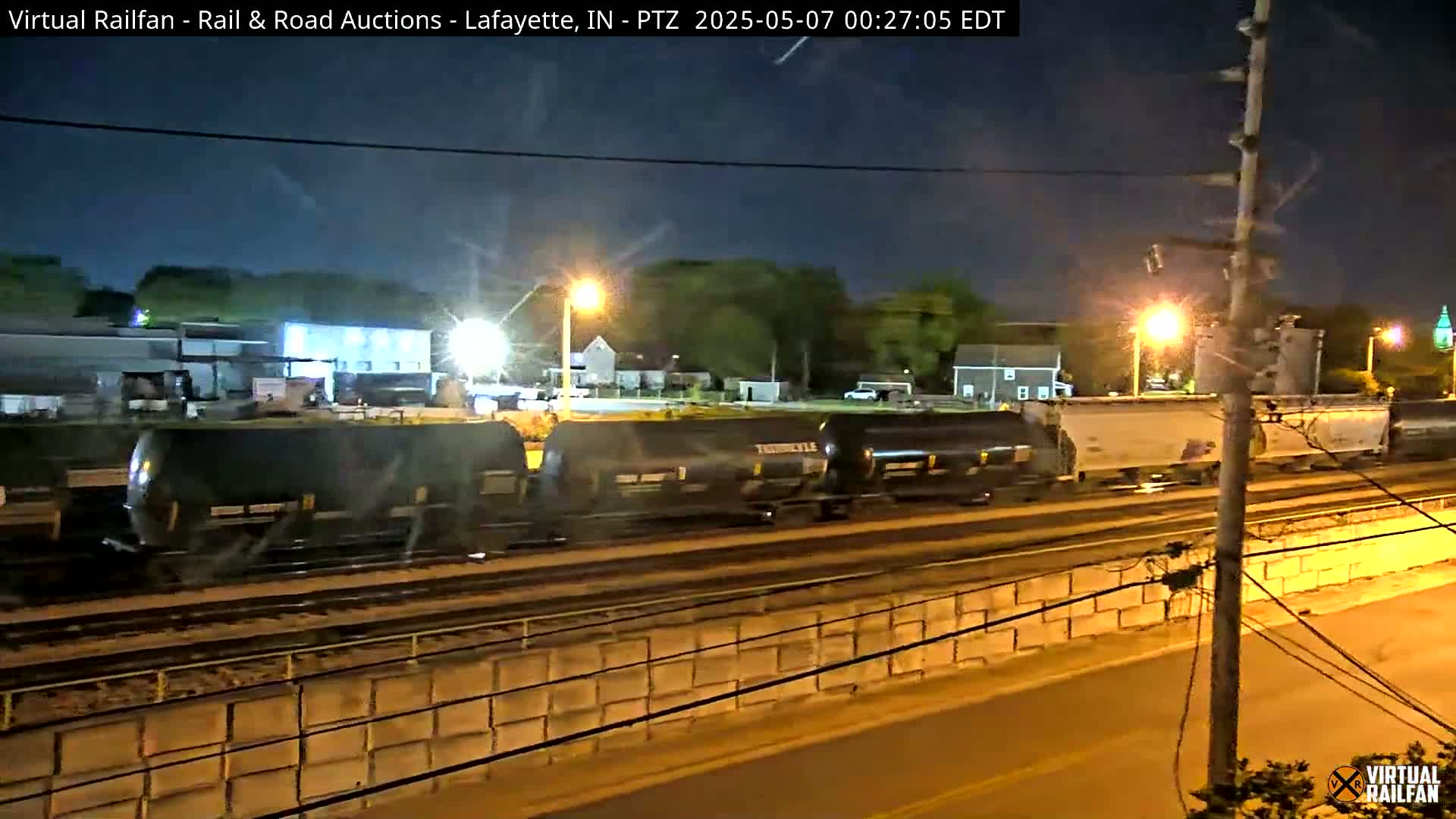 At night, a long freight train moves along railroad tracks, passing under streetlights beside a low concrete wall.