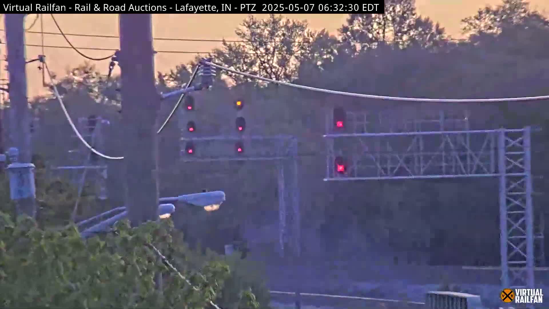 A hazy sunrise illuminates a railroad crossing with multiple red signal lights activated, surrounded by trees and utility poles.