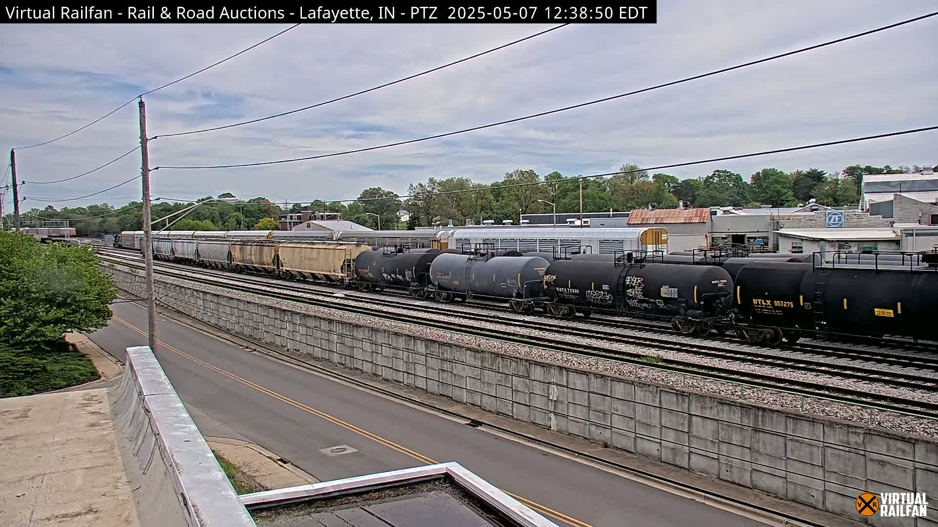 A long freight train, consisting of various cars including tank cars and covered hopper cars, is seen on a railway track next to a road under a partly cloudy sky.
