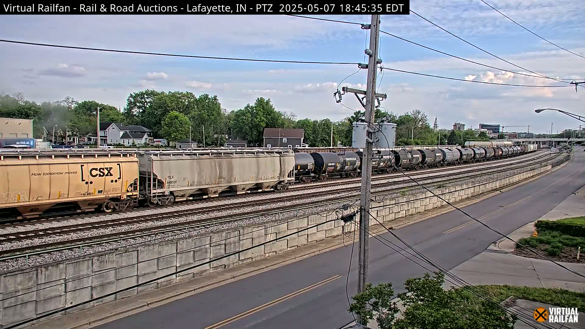 A long freight train, primarily consisting of tanker cars, moves along a railway track beside a road under a partly cloudy sky.