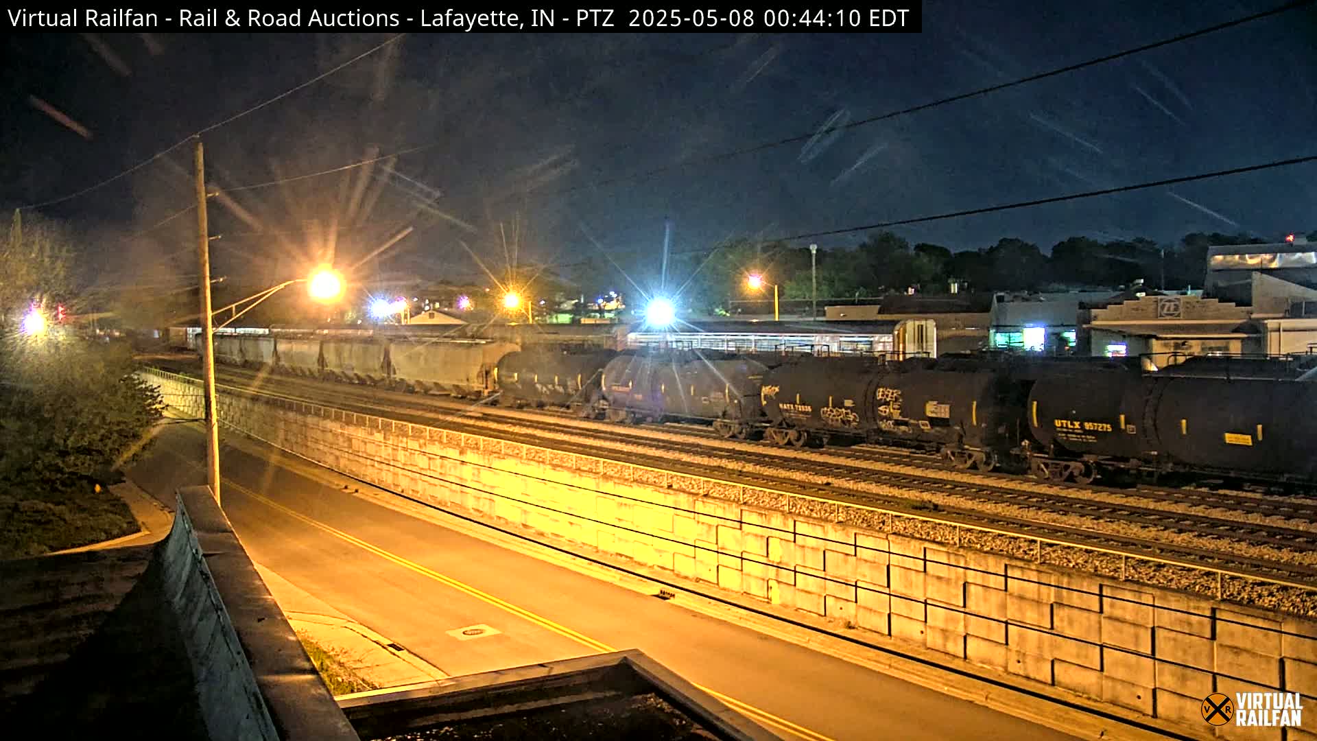 At night, a long freight train sits on railroad tracks next to a concrete retaining wall beside a road.