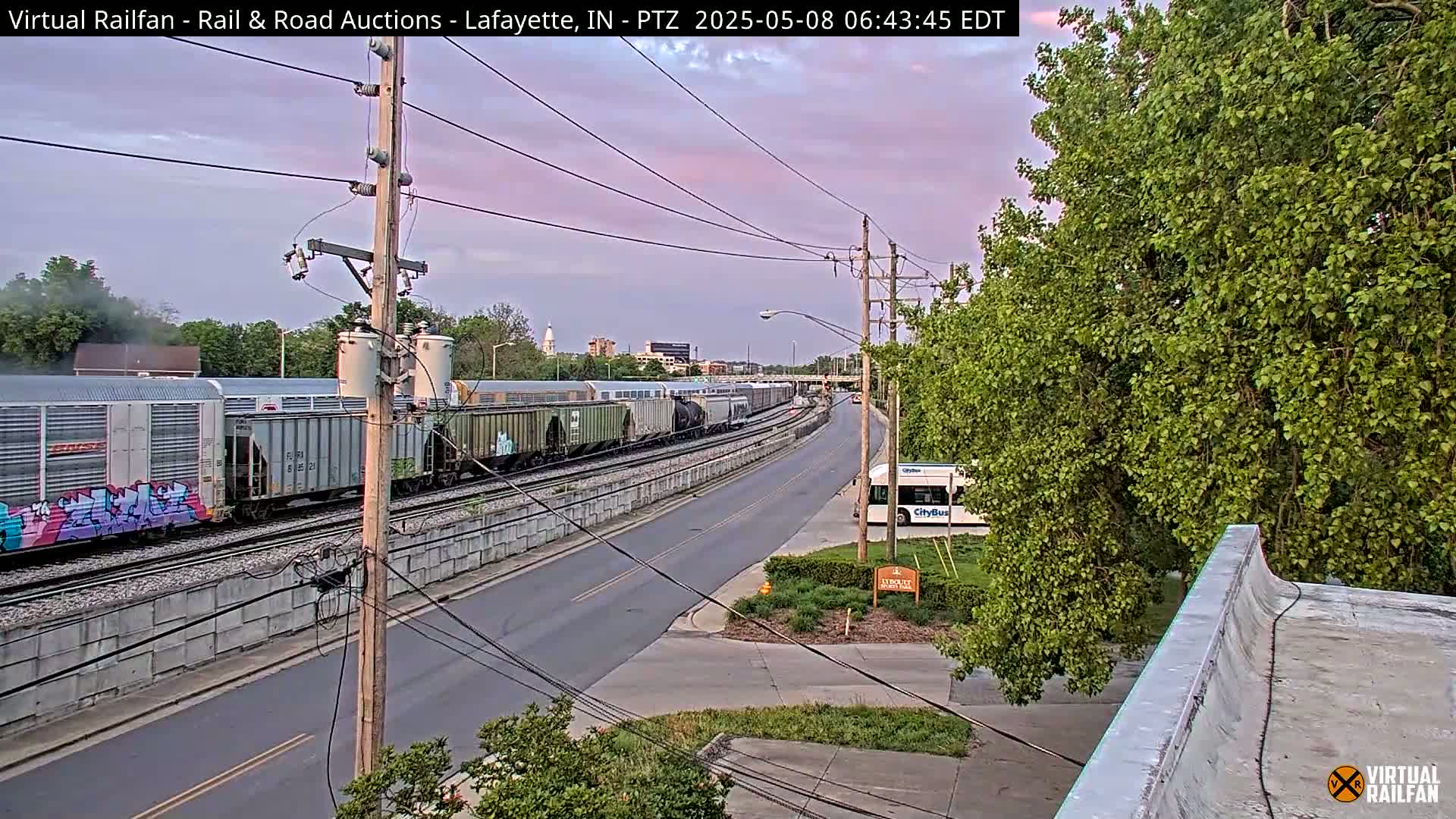 A long freight train passes by a city street under a pale pink twilight sky.