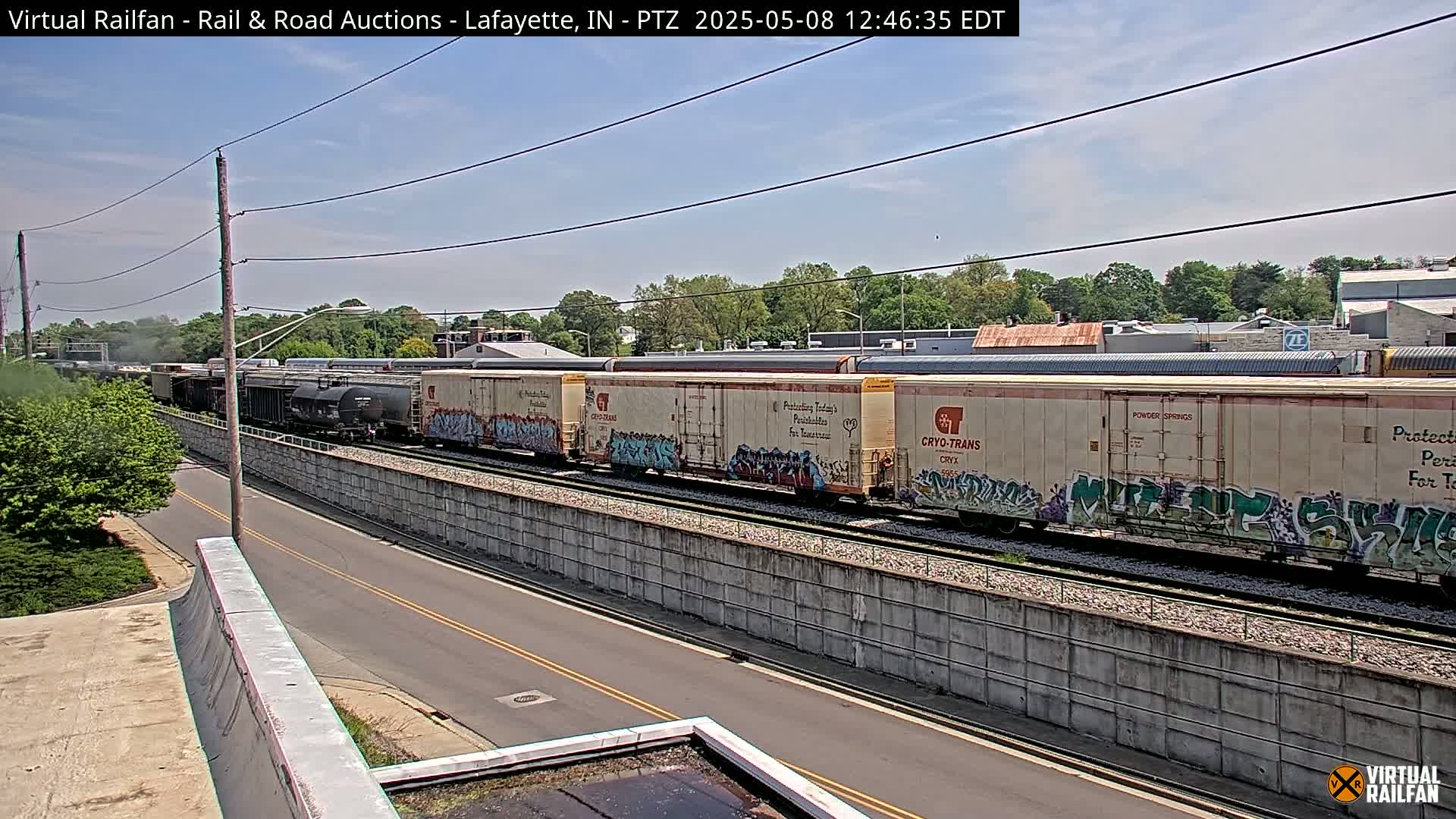 A long freight train with graffiti-covered cars moves along a track beside a road under a mostly sunny sky.