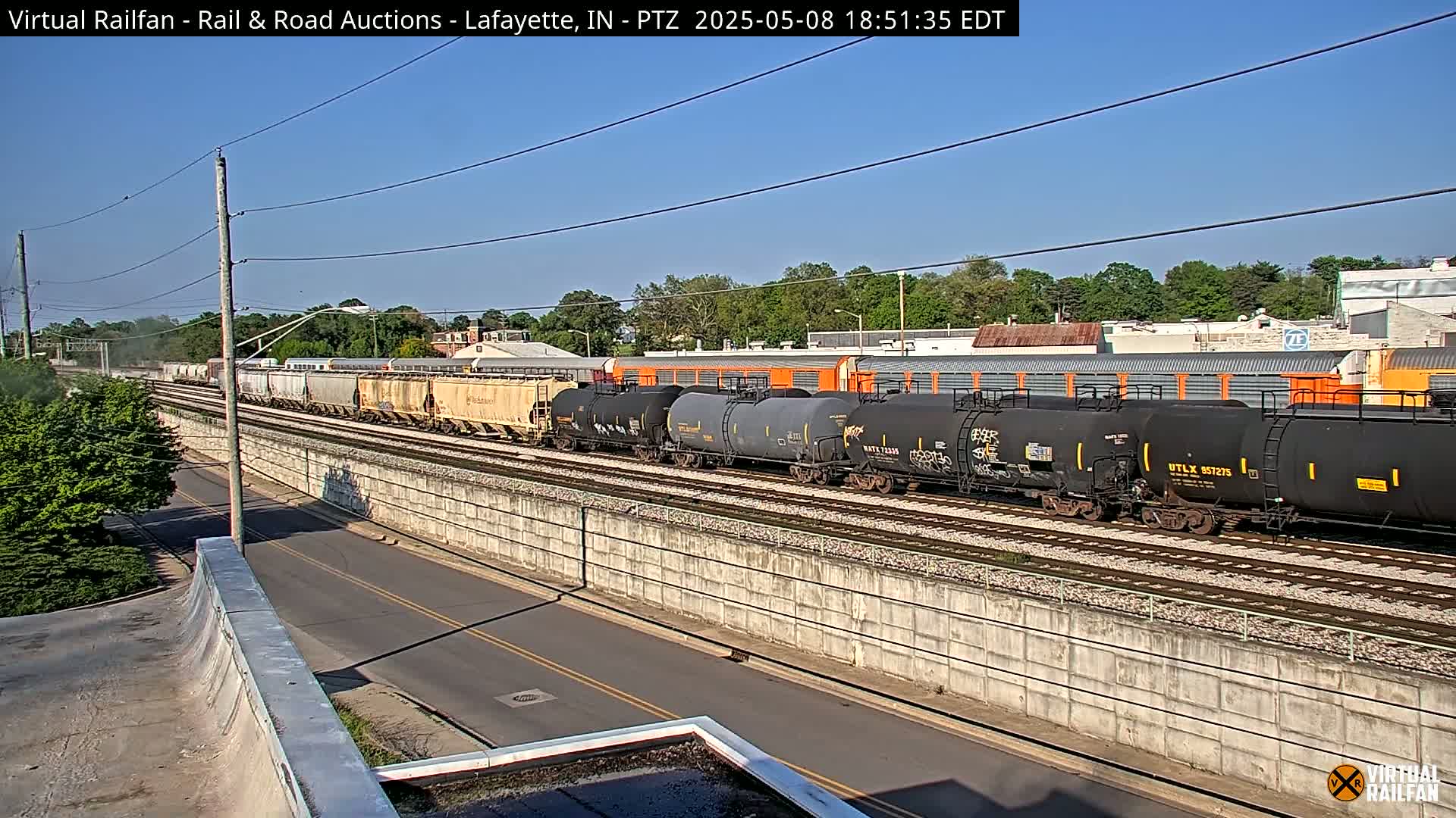 A long freight train, consisting of both boxcars and tank cars, is seen traveling along railroad tracks on a sunny day next to a road.