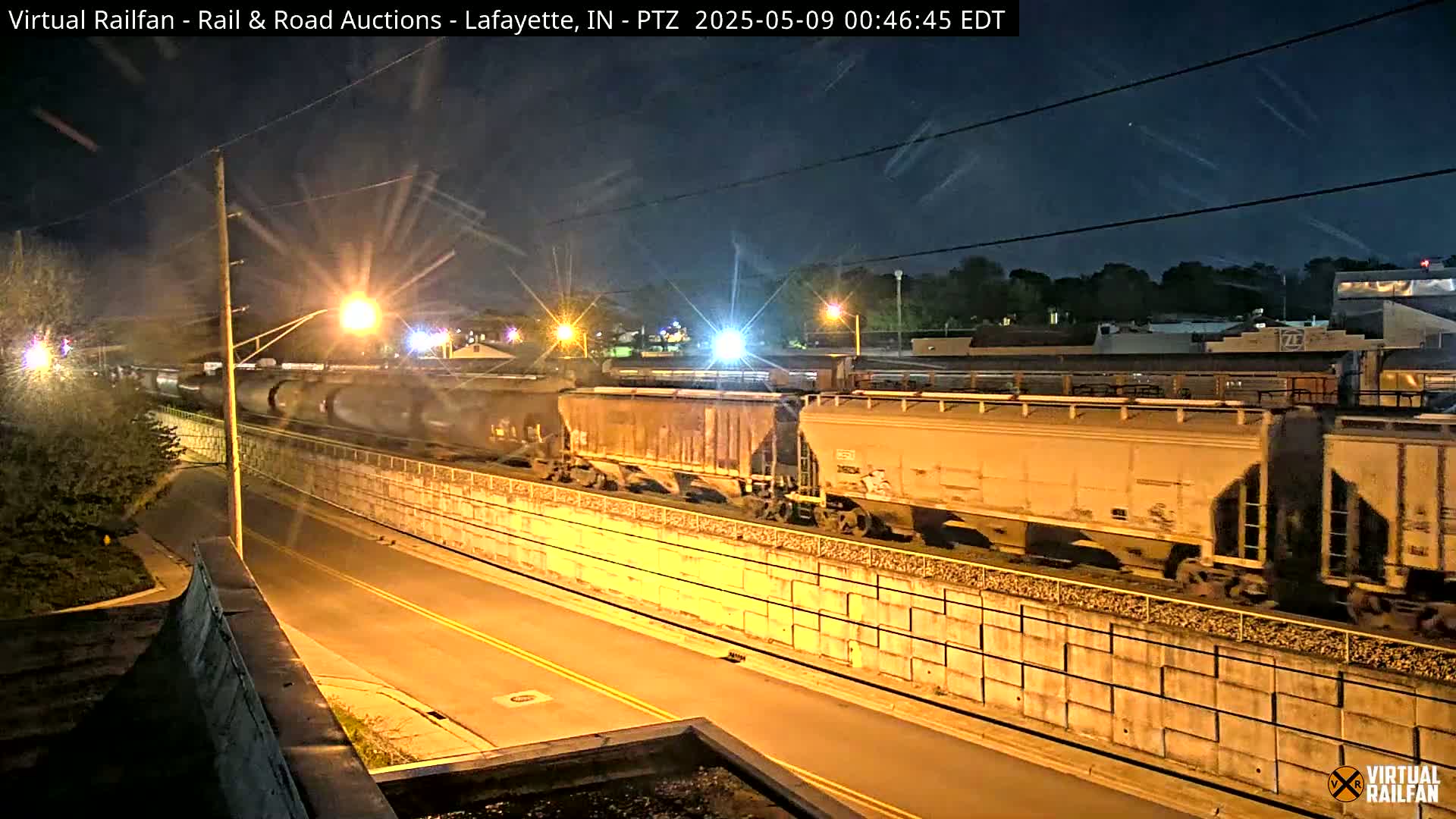 At night, a freight train passes behind a concrete retaining wall next to a road lit by streetlights.