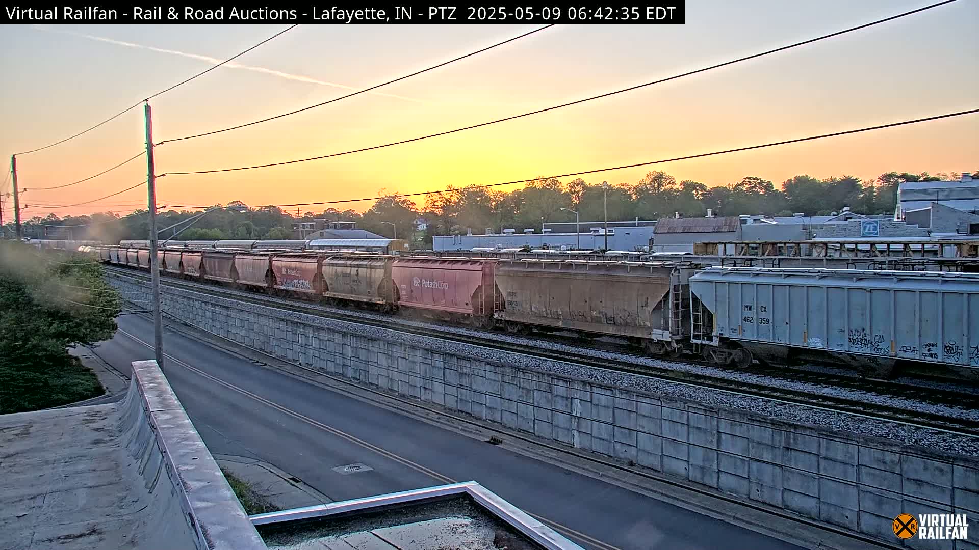 A long freight train passes by at sunrise next to a road and a tall concrete wall.