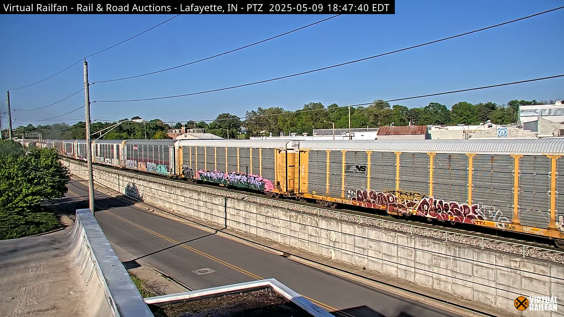 A long freight train with graffiti-covered cars travels along railroad tracks next to a road on a sunny day.