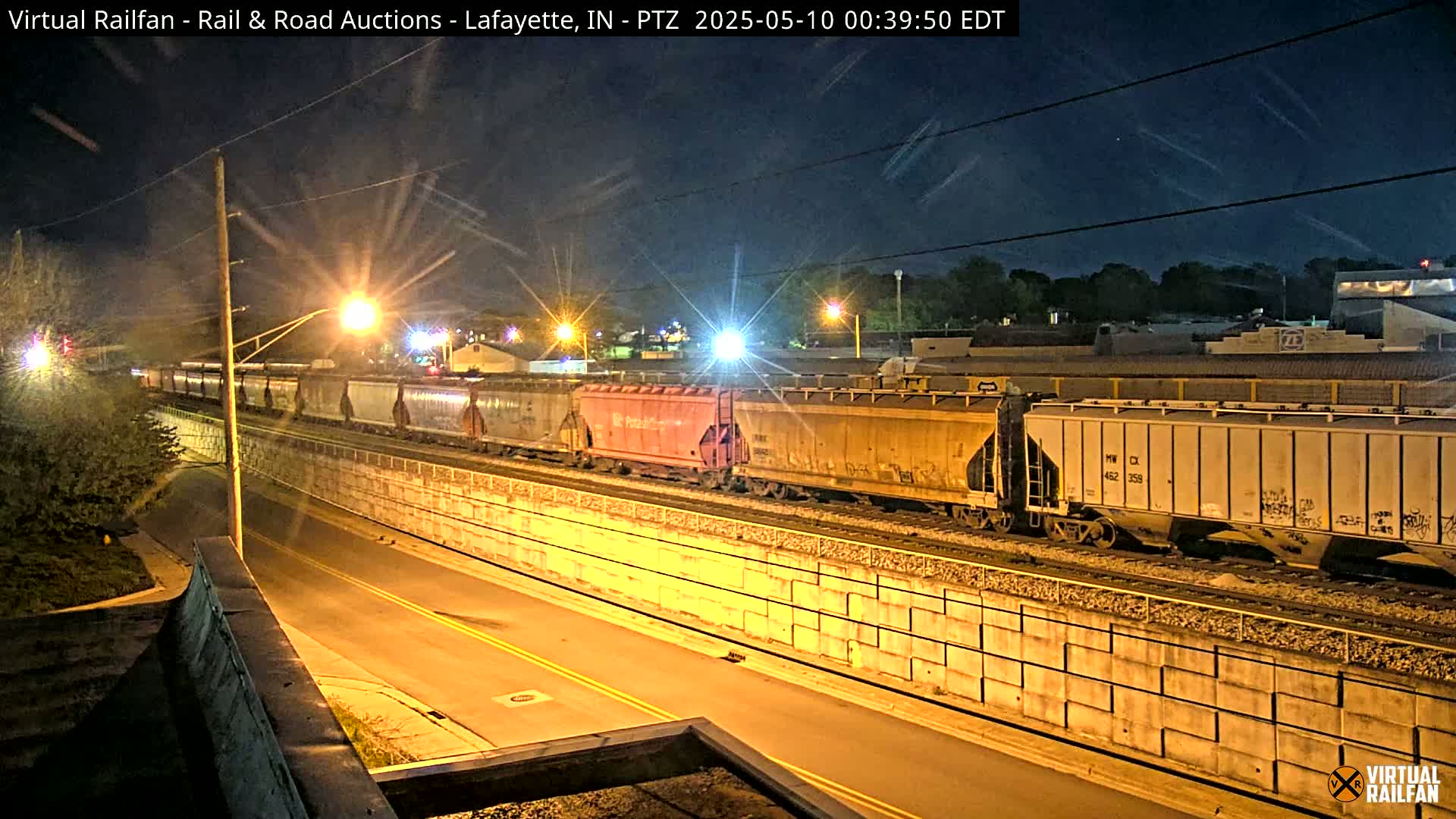 At night, a long freight train travels along a track beside a roadway, illuminated by streetlights.