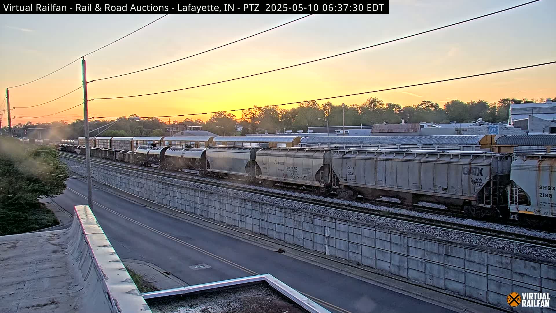 A long freight train passes by during a sunrise with a clear, pale orange sky.
