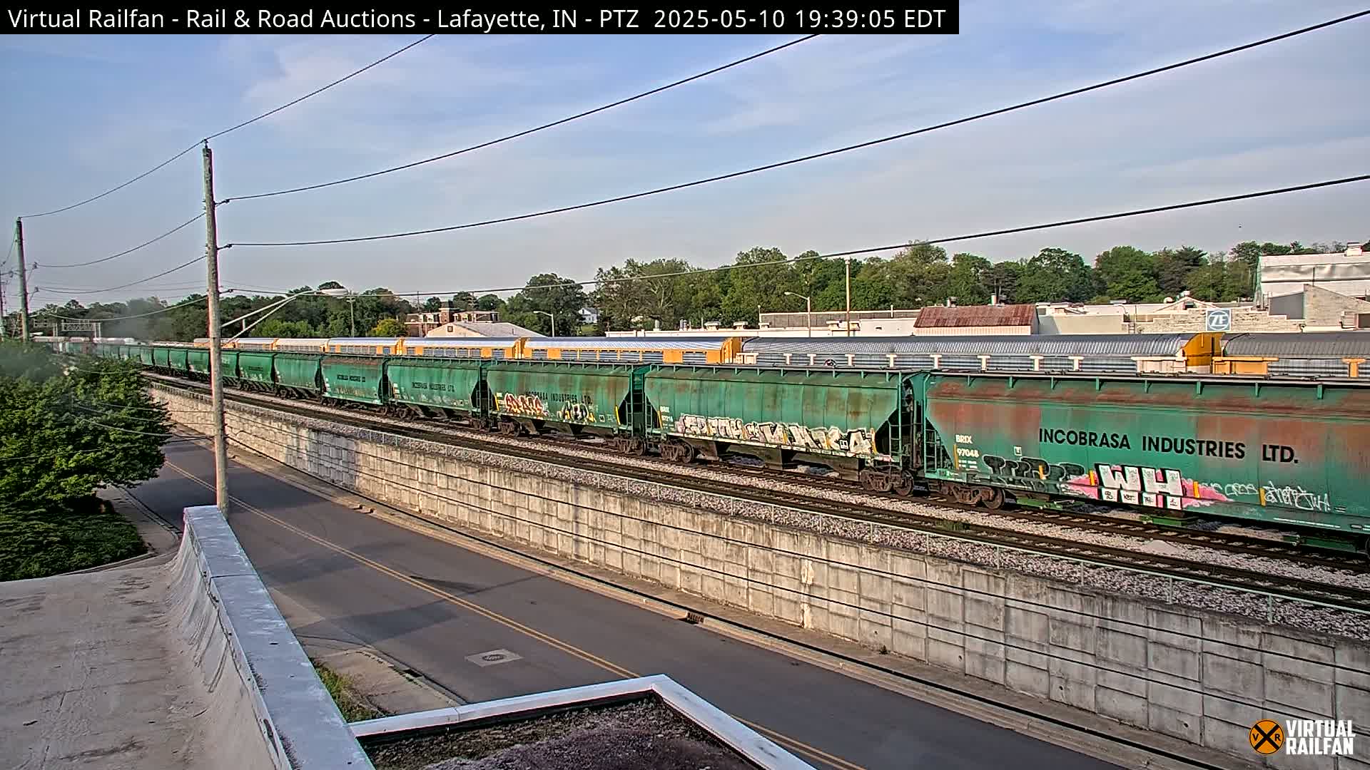 A long freight train, some cars of which are green and covered in graffiti, travels along railroad tracks under a partly cloudy sky.