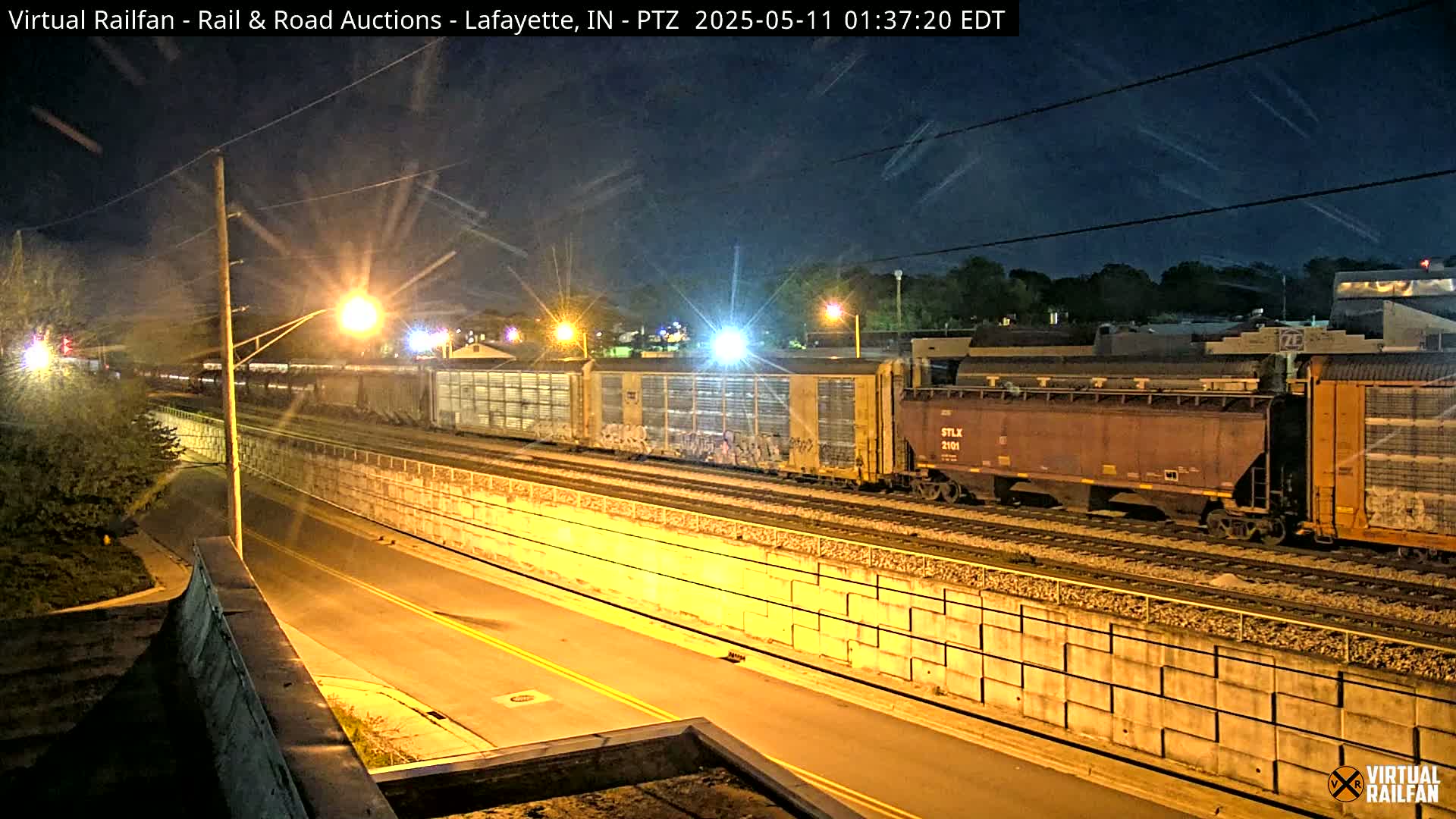 At night, a freight train sits on railroad tracks next to a road, separated by a concrete barrier, under a clear, dark sky.