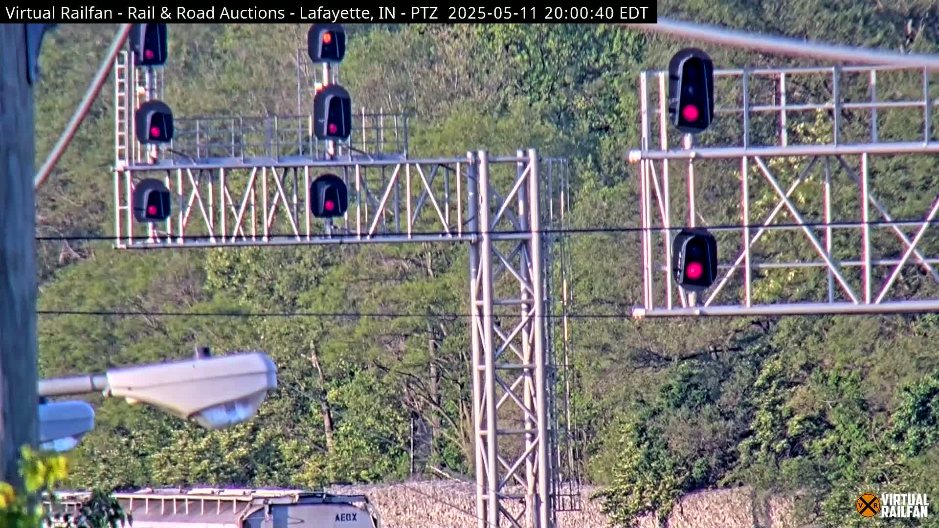 Multiple red railroad signals are shown against a backdrop of green trees and a partially visible train car under a daytime sky.