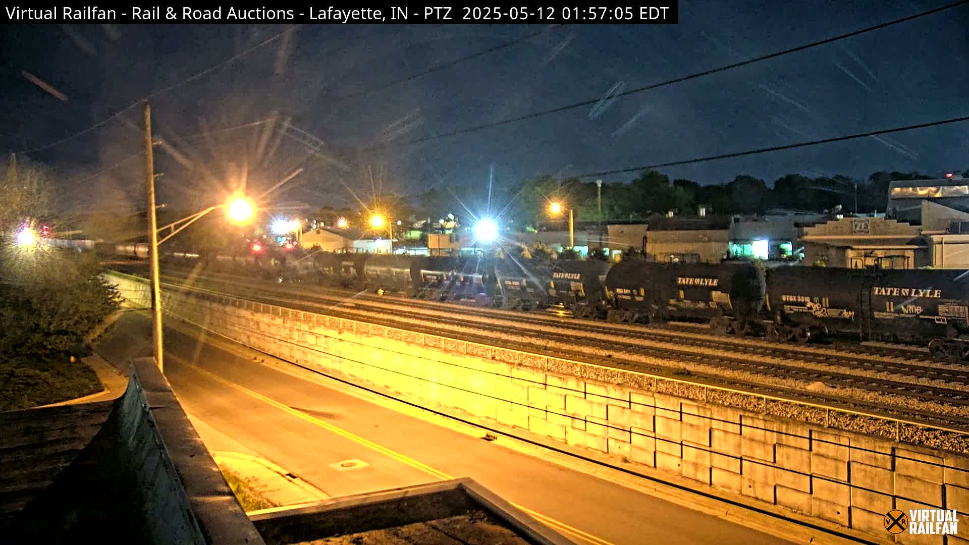 At night, a long freight train sits on railroad tracks beside a roadway bordered by a high retaining wall, under a mostly clear sky.