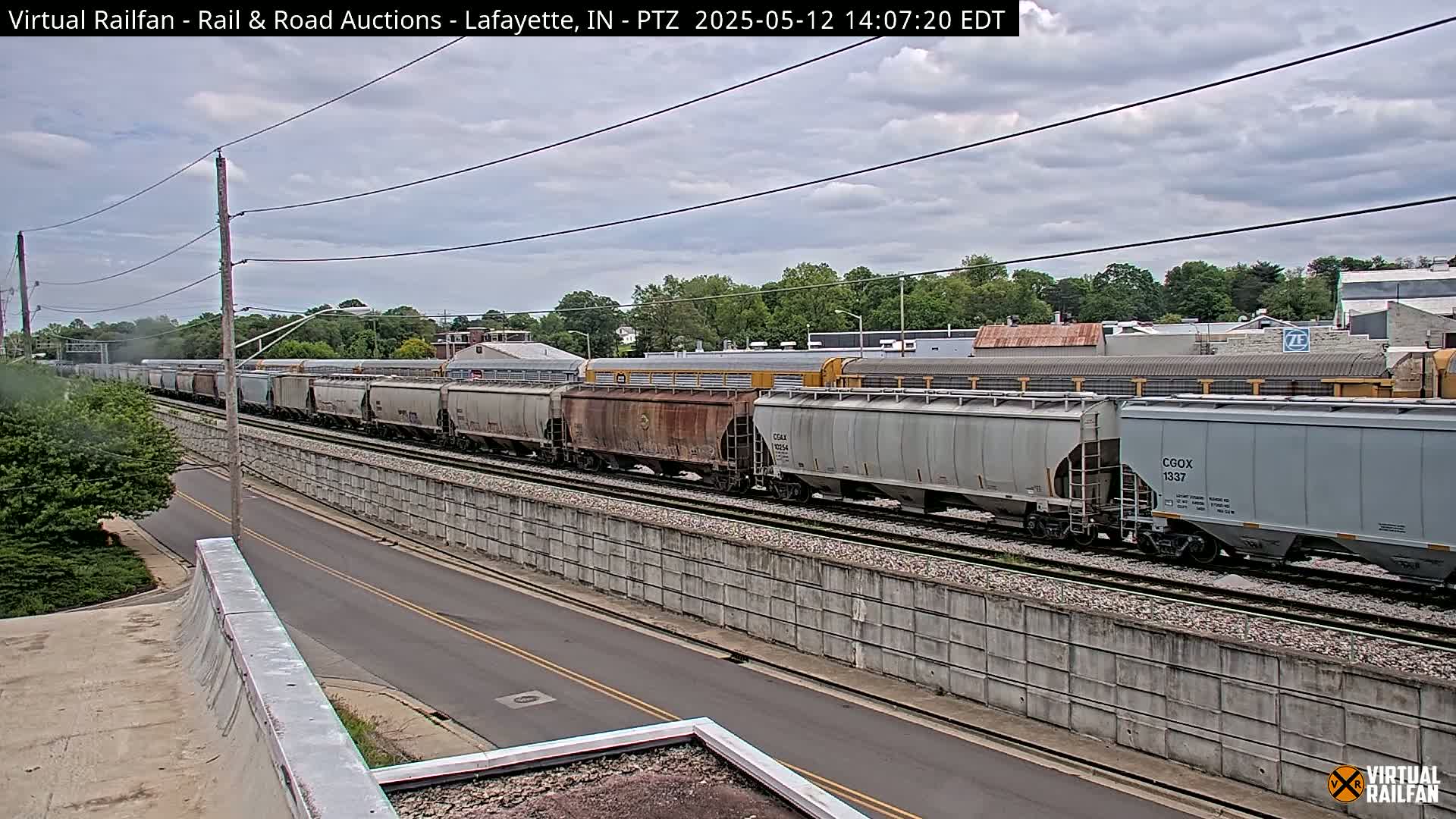 A long freight train passes by under a partly cloudy sky next to a road and a concrete wall.