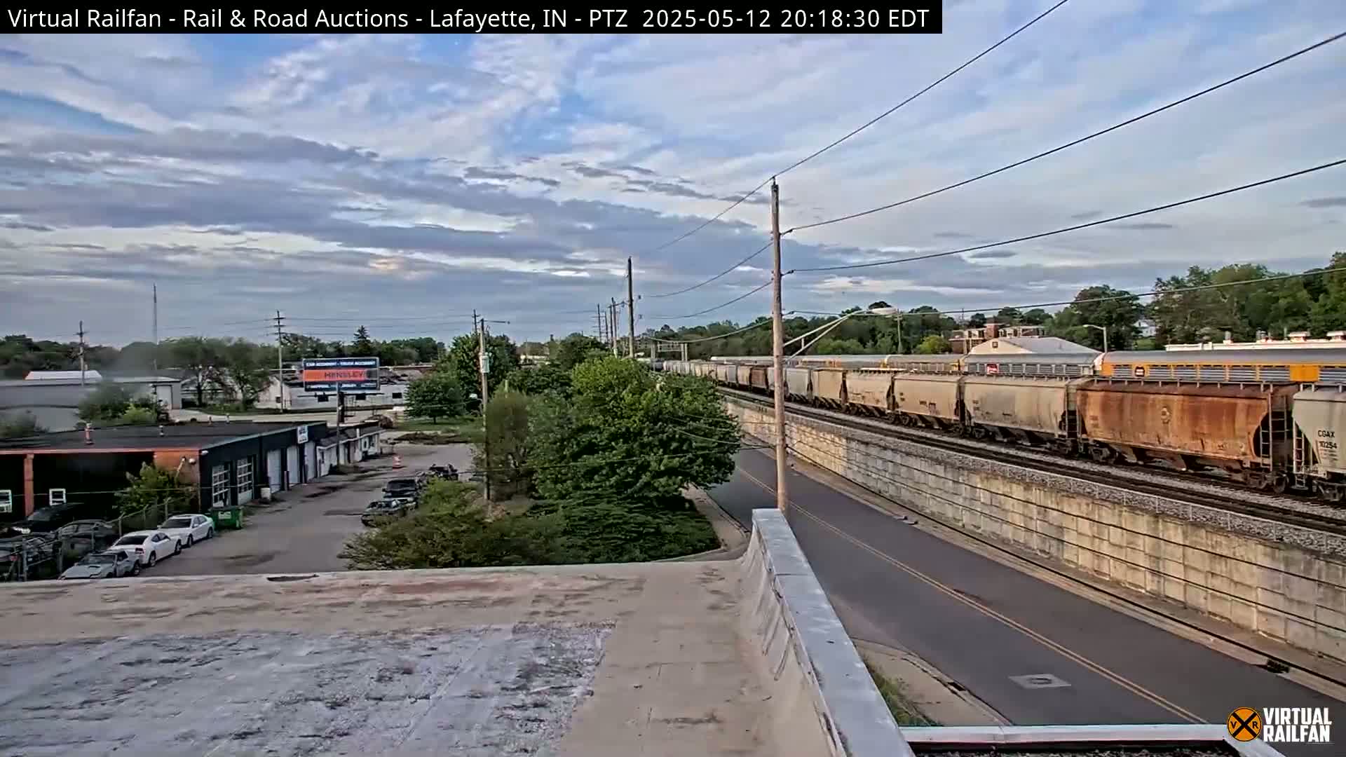 A long freight train passes by under a partly cloudy evening sky, next to a road and various buildings including a car repair shop.