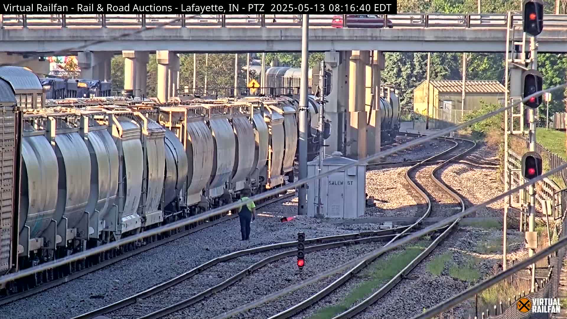 A long freight train of covered hopper cars sits on a rail line under a bridge on a sunny day.