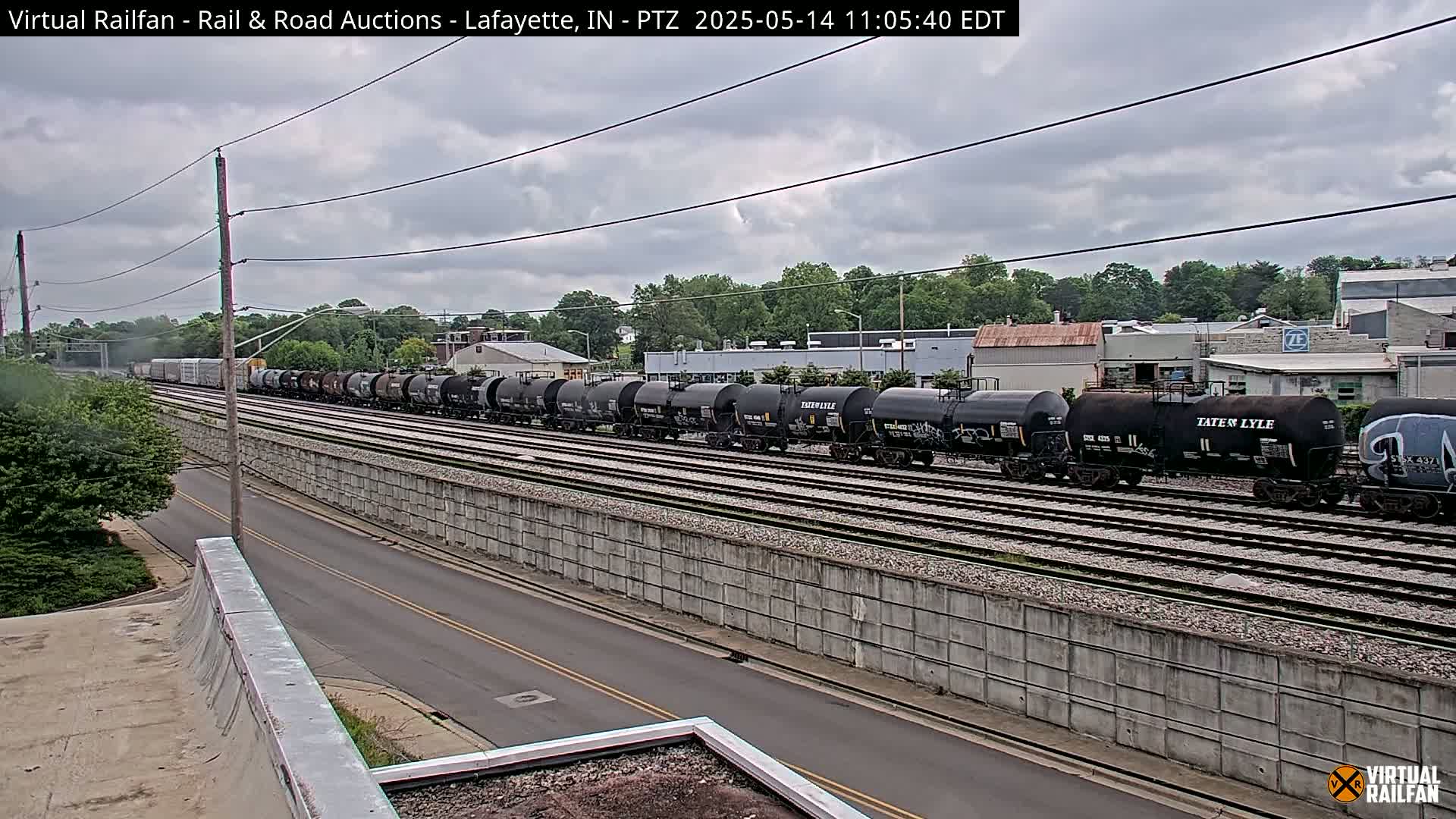 A long freight train of tanker cars sits on railroad tracks next to a road under an overcast sky.