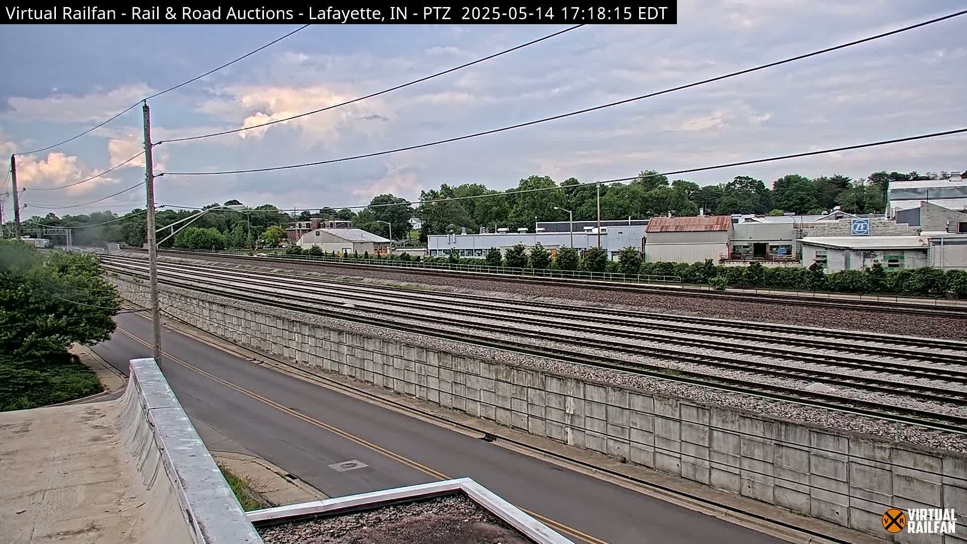 A high-angle view shows a section of railroad tracks next to a road, with a concrete sound barrier separating them under a partly cloudy sky.