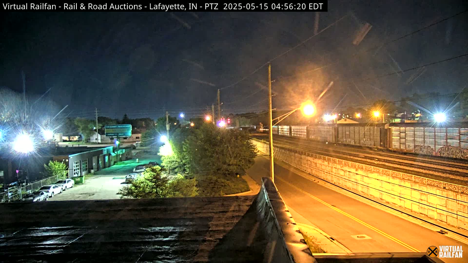 At night, a long freight train passes under a streetlight-lit overpass beside a road, with cars parked in a lot visible in the distance.
