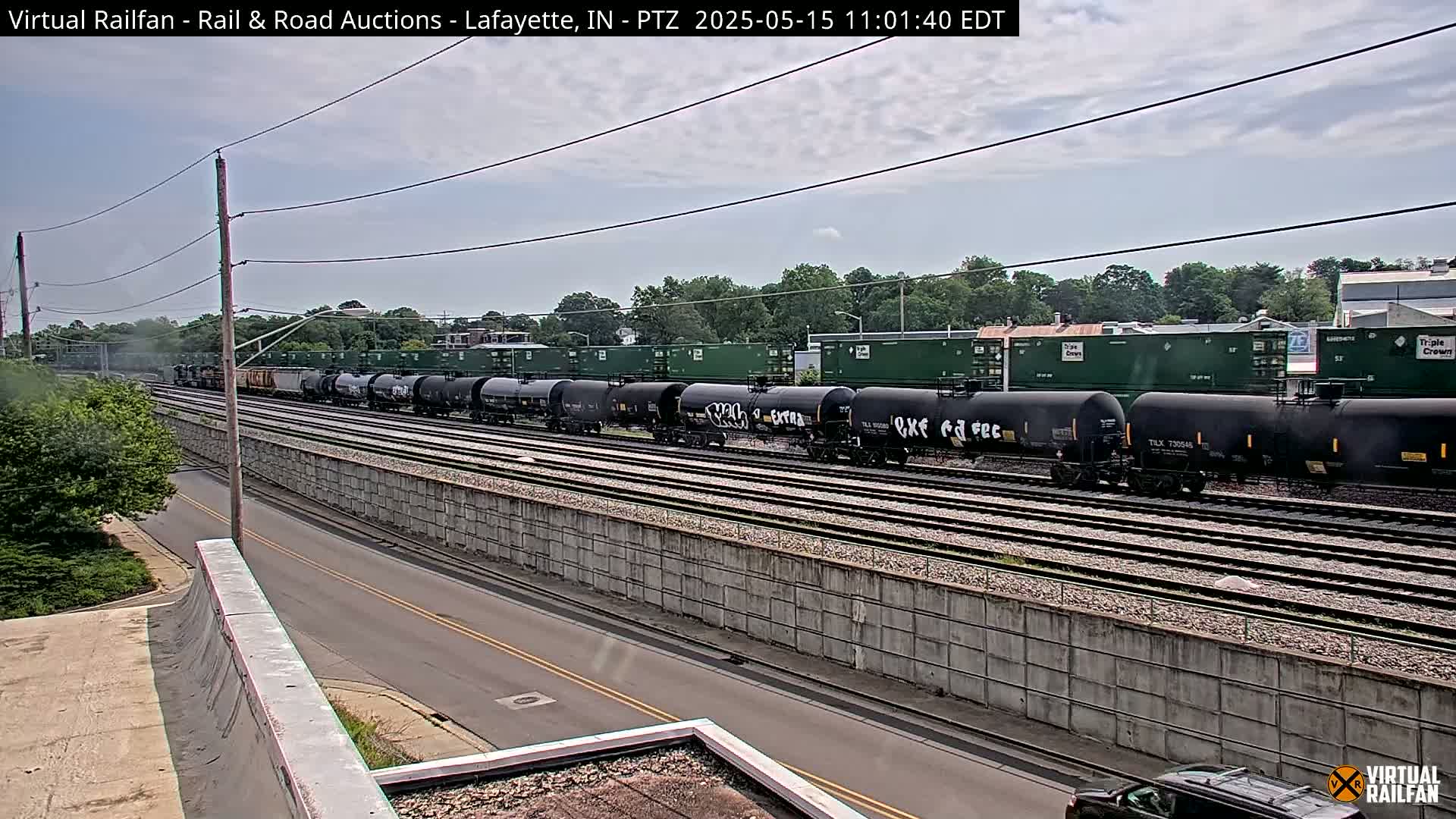 A long freight train of tanker cars sits on a railway track next to a road, under a partly cloudy sky.