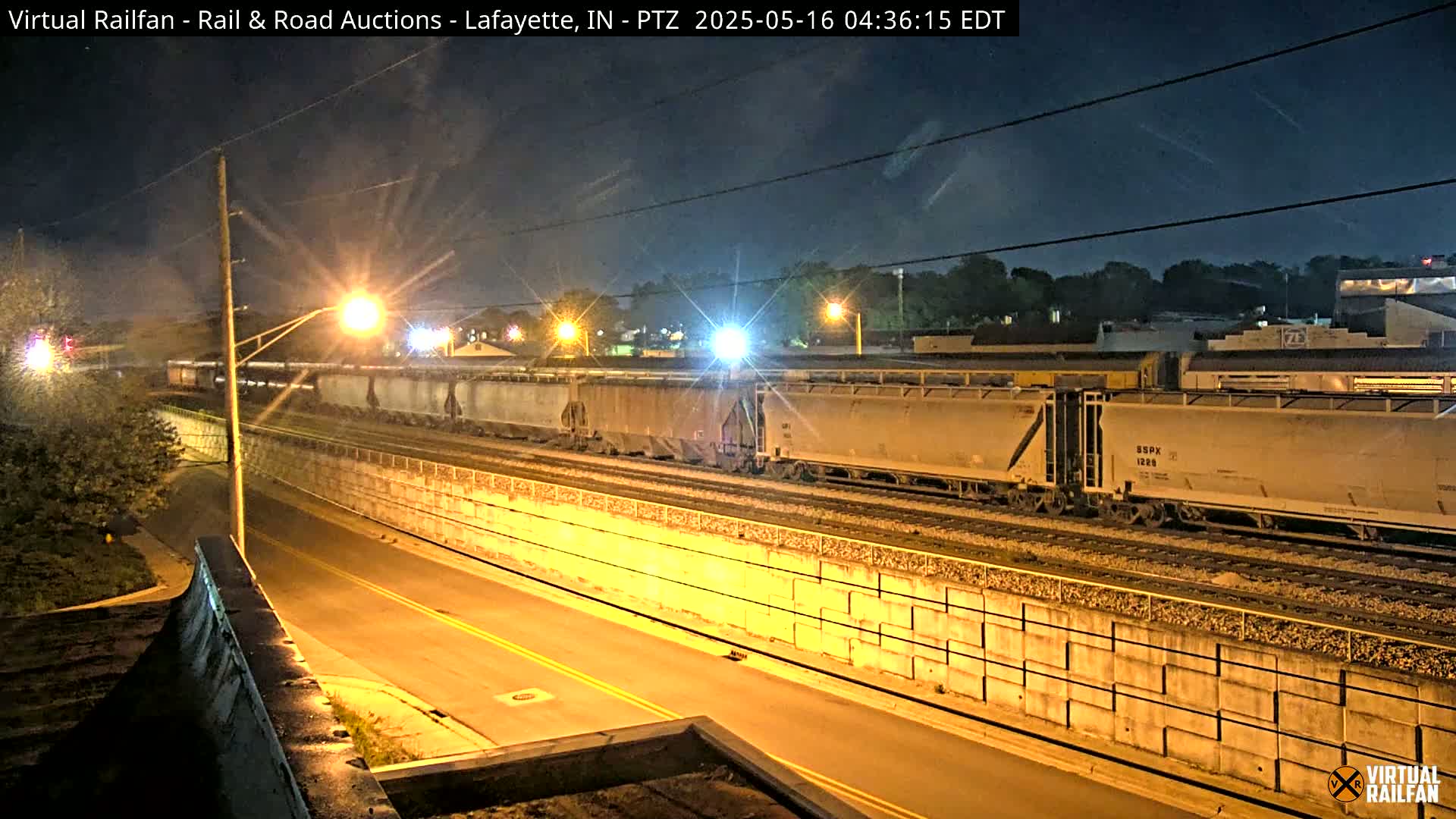 A long freight train travels along railroad tracks at night under a clear, dark sky illuminated by streetlights.